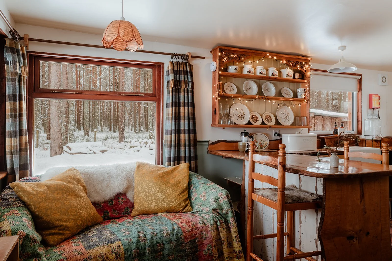 Cozy living room with a large window revealing a snowy outdoor scene. A couch with colorful patchwork fabric and two gold pillows sits by the window, covered with a white furry throw. To the right, a wooden kitchen counter with a barstool, plates, an