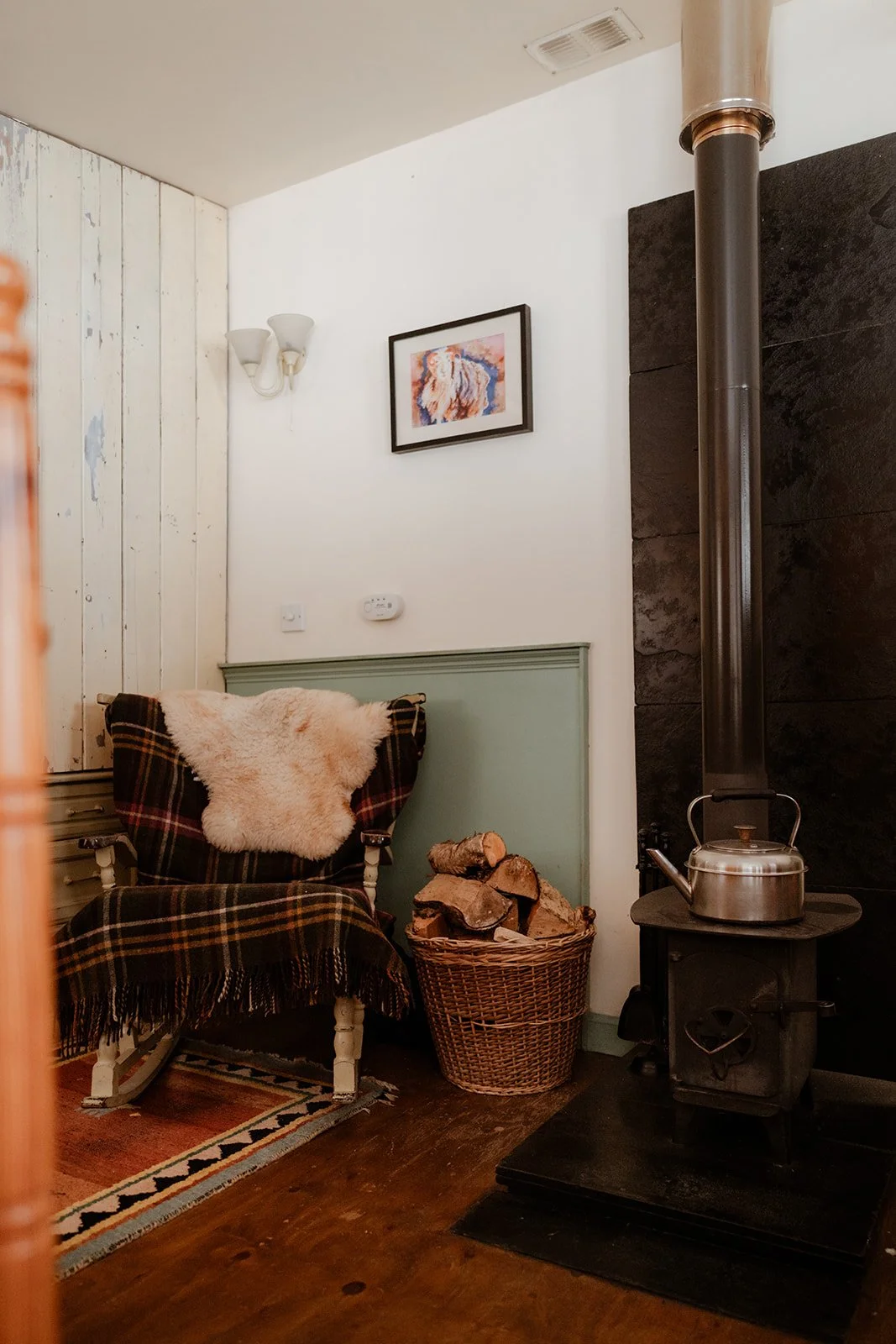 Cozy corner with a wooden armchair draped with a plaid blanket and sheepskin, next to a basket of firewood, with a wood stove and kettle, and a framed picture on the wall.