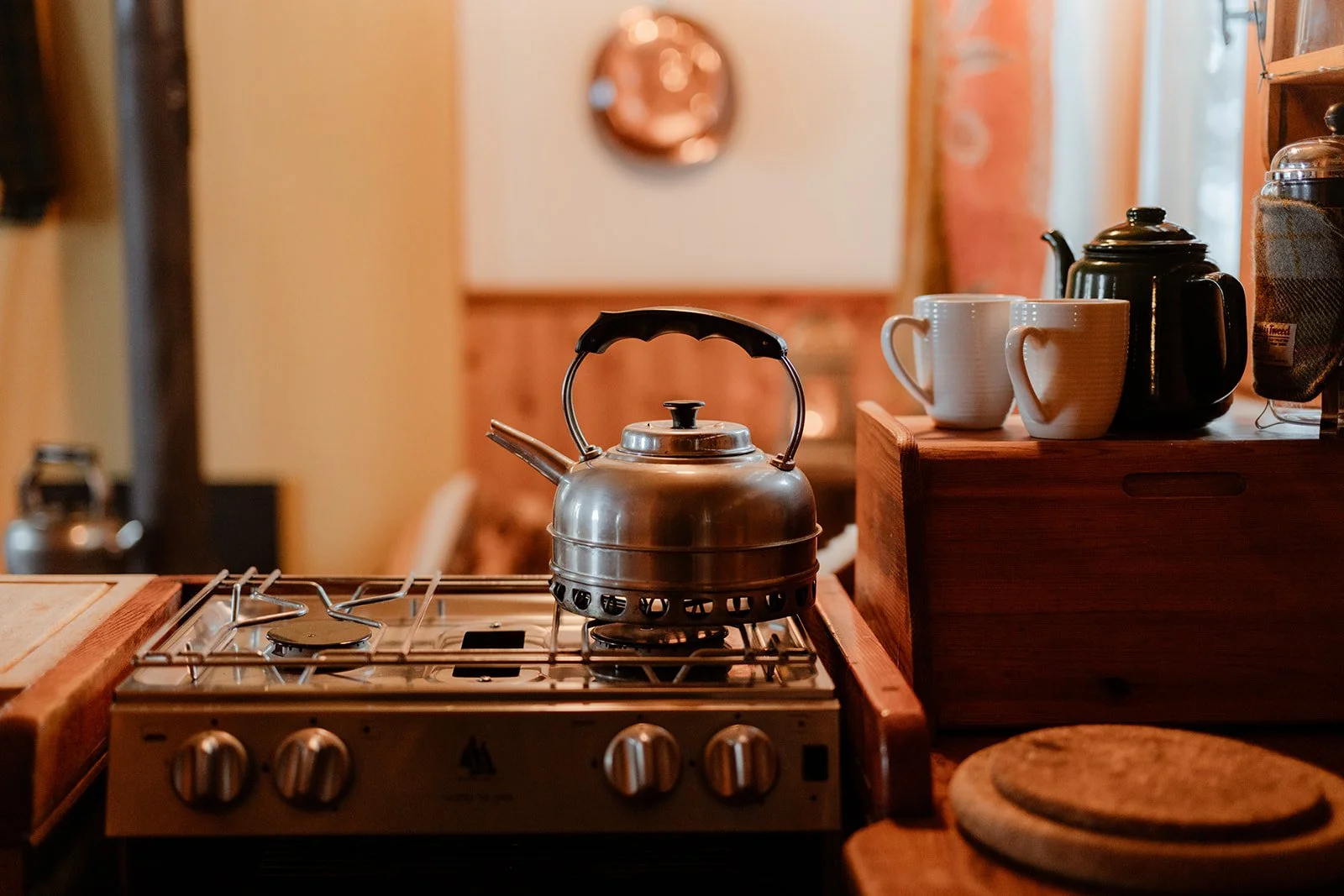 A kitchen stove with a silver kettle on top, next to wooden countertop with two white mugs, a black teapot, and a coffee jar.