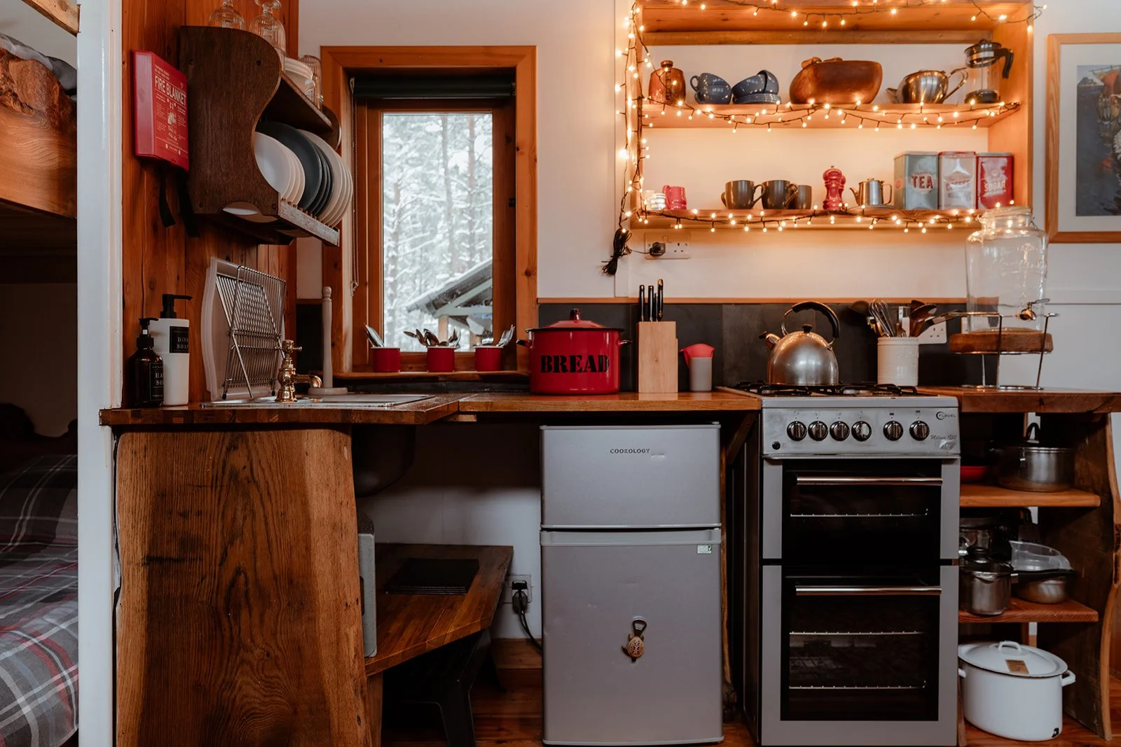 A cozy kitchen with wooden cabinetry and shelves, string lights, a small window showing snow outside, a red bread box on the counter, a stove, a mini refrigerator, and various kitchenware and decor.
