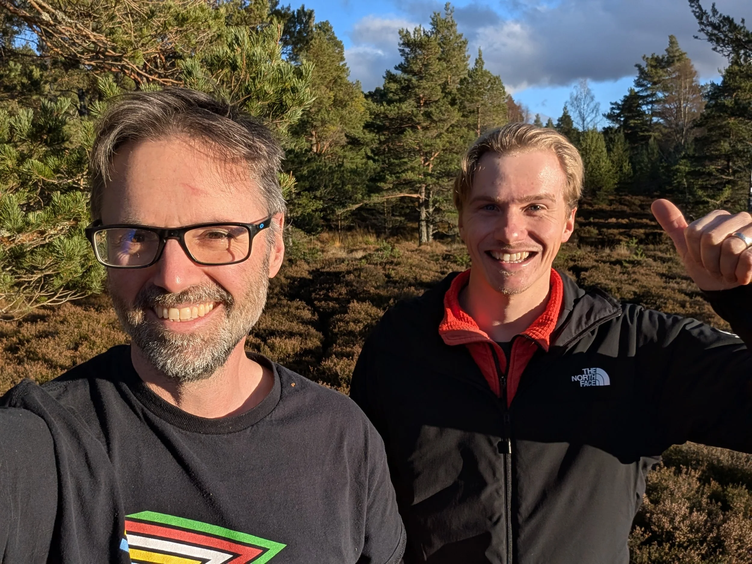 Two smiling men outdoors in a wooded area, taking a selfie, with trees and blue sky in the background.