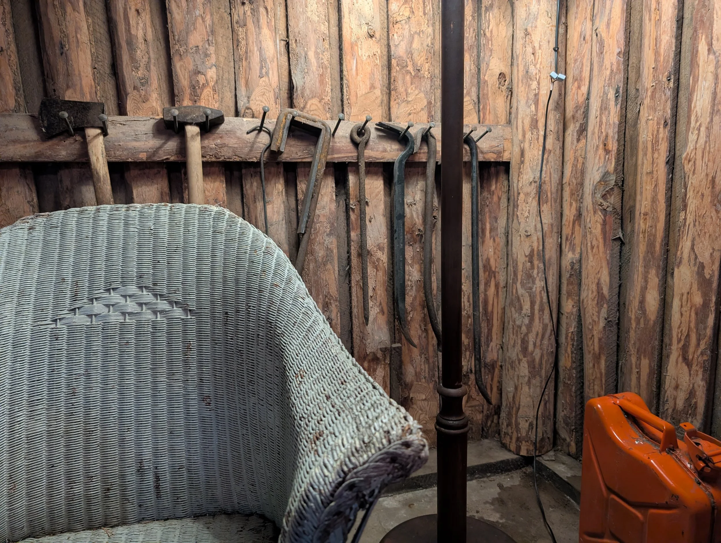 A rustic workshop corner with wooden walls, hanging antique tools, a wicker chair, an orange fuel container, and a black lamp standing on the floor.