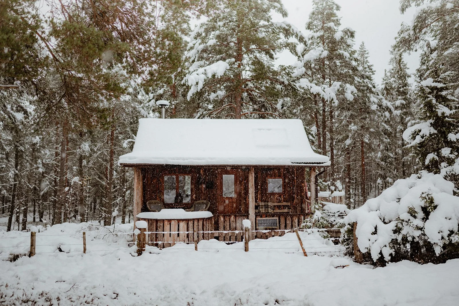 The Woodman's Hut covered in snow - an off-grid cabin surrounded by scots pine forest with a wood-burning stove, whisky barrel in front and outdoor bucket shower to one side