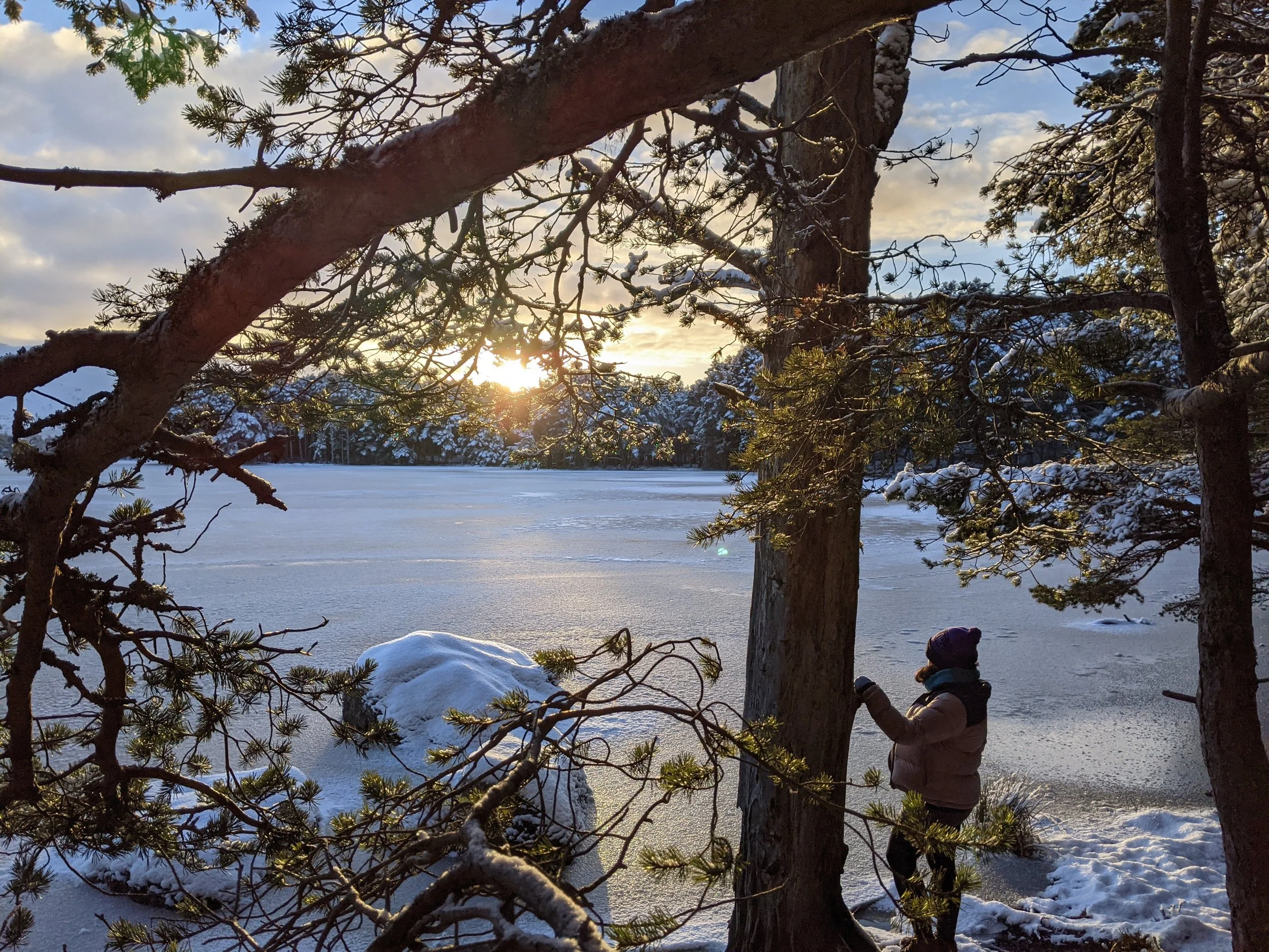 a frozen loch, with the sun setting and a women looking out over the frozen waters.