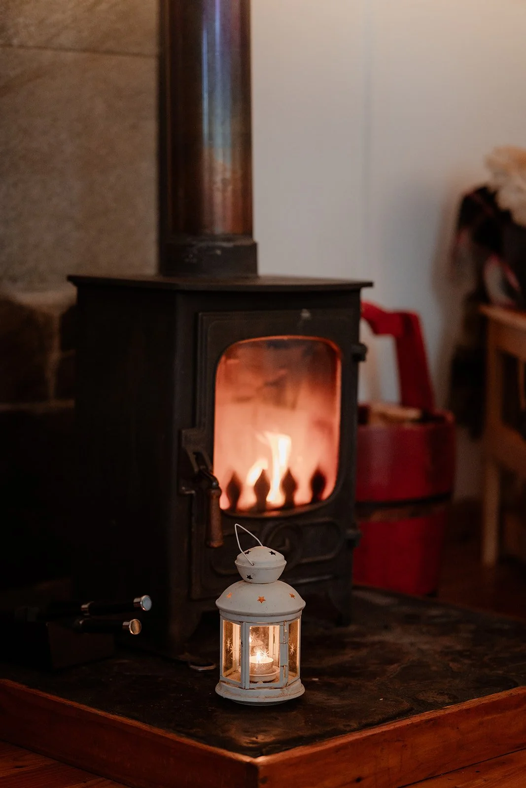 A lit stove with a glowing fire inside, placed on a wooden surface, with a small white lantern and candle in front, creating a cozy ambiance.