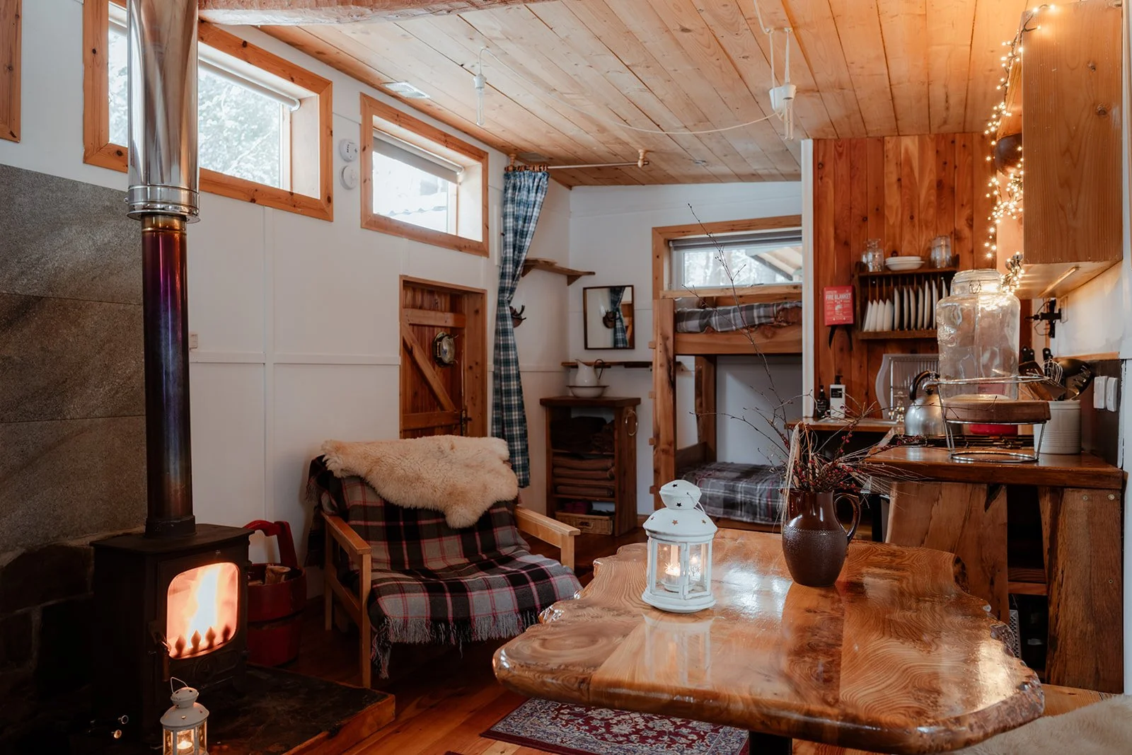 Cozy cabin interior with wood-paneled ceiling, windows, a wood stove with a lit fire, a plaid armchair with a sheepskin throw, a wooden table with a lantern, and bunk beds in the background.