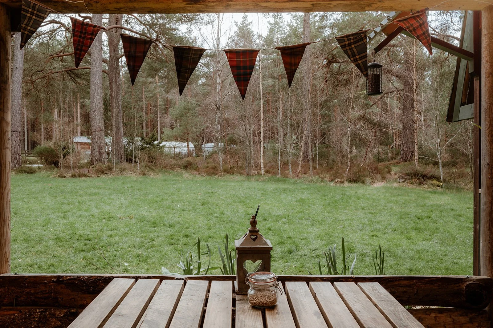 View from a porch looking out over a grassy backyard with trees in the distance. The porch has a wooden table, a lantern, and a glass jar on it, with bunting flags hanging overhead.
