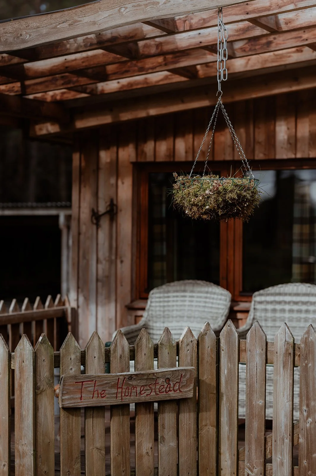 The image depicts a rustic wooden porch with a fence in the foreground that has a small wooden sign saying "The Homestead". Behind the fence, there are two wicker chairs, a window with a wooden frame, and a hanging plant composed of dried herbs or fl