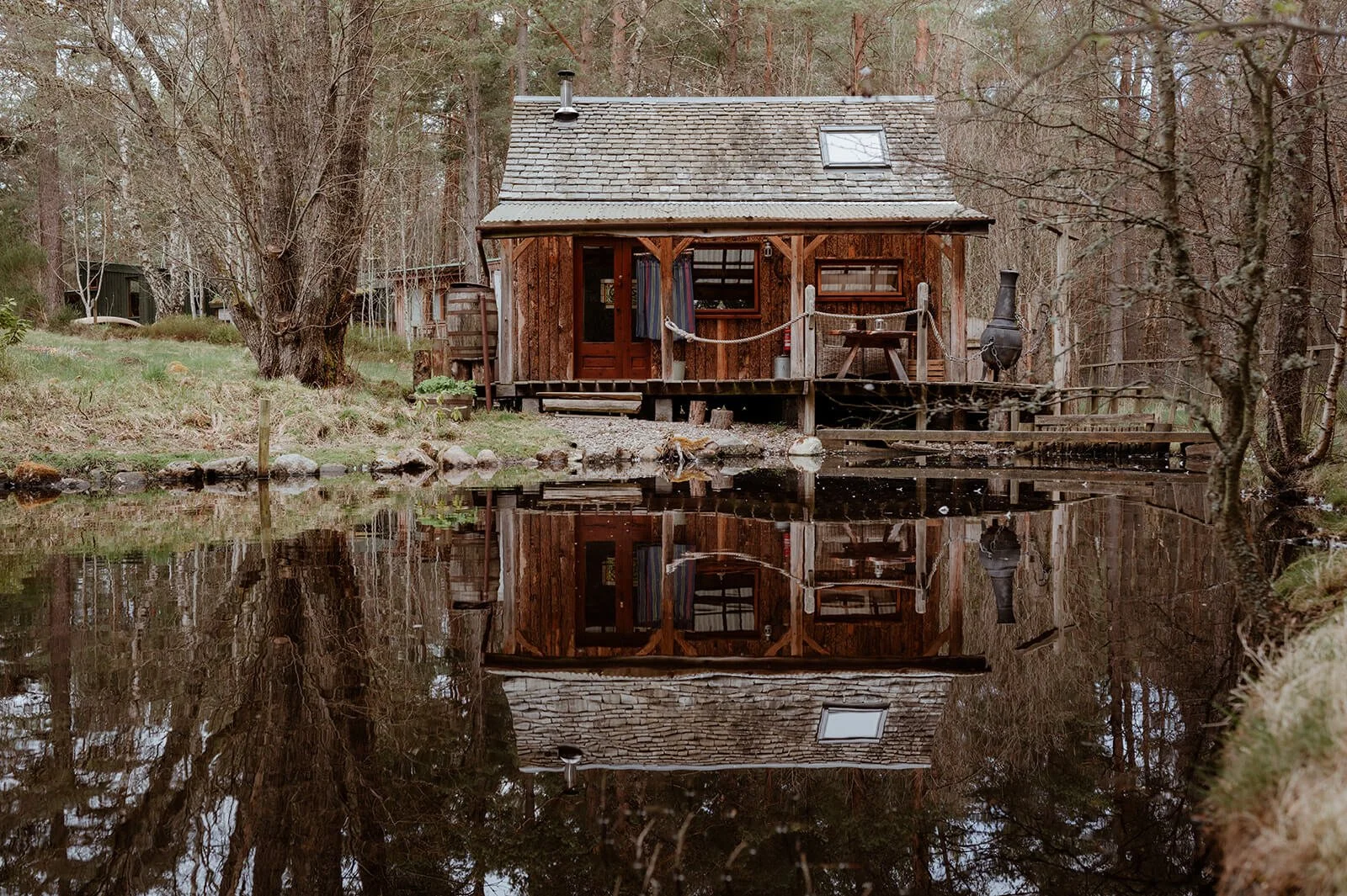 A forest off-grid hut relfected in the stil waters of a pond surrounded by pine trees.