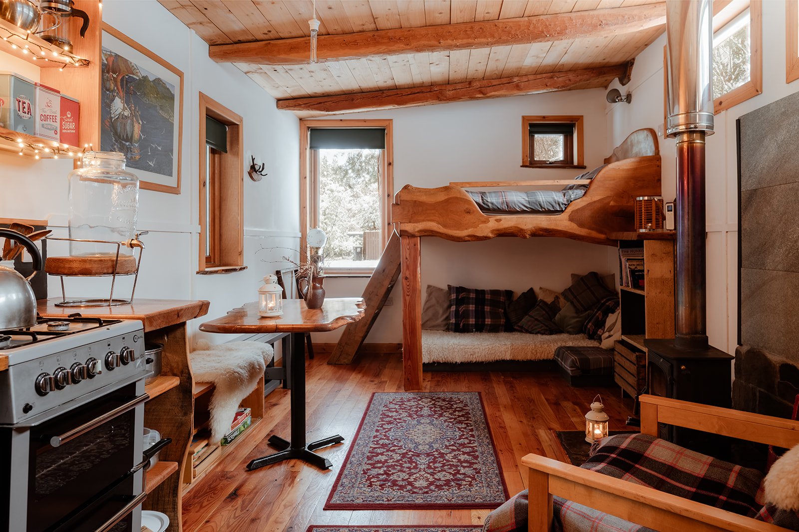 Cozy cabin interior with a wooden loft bed, a small table, a stove, and decor in a rustic style.
