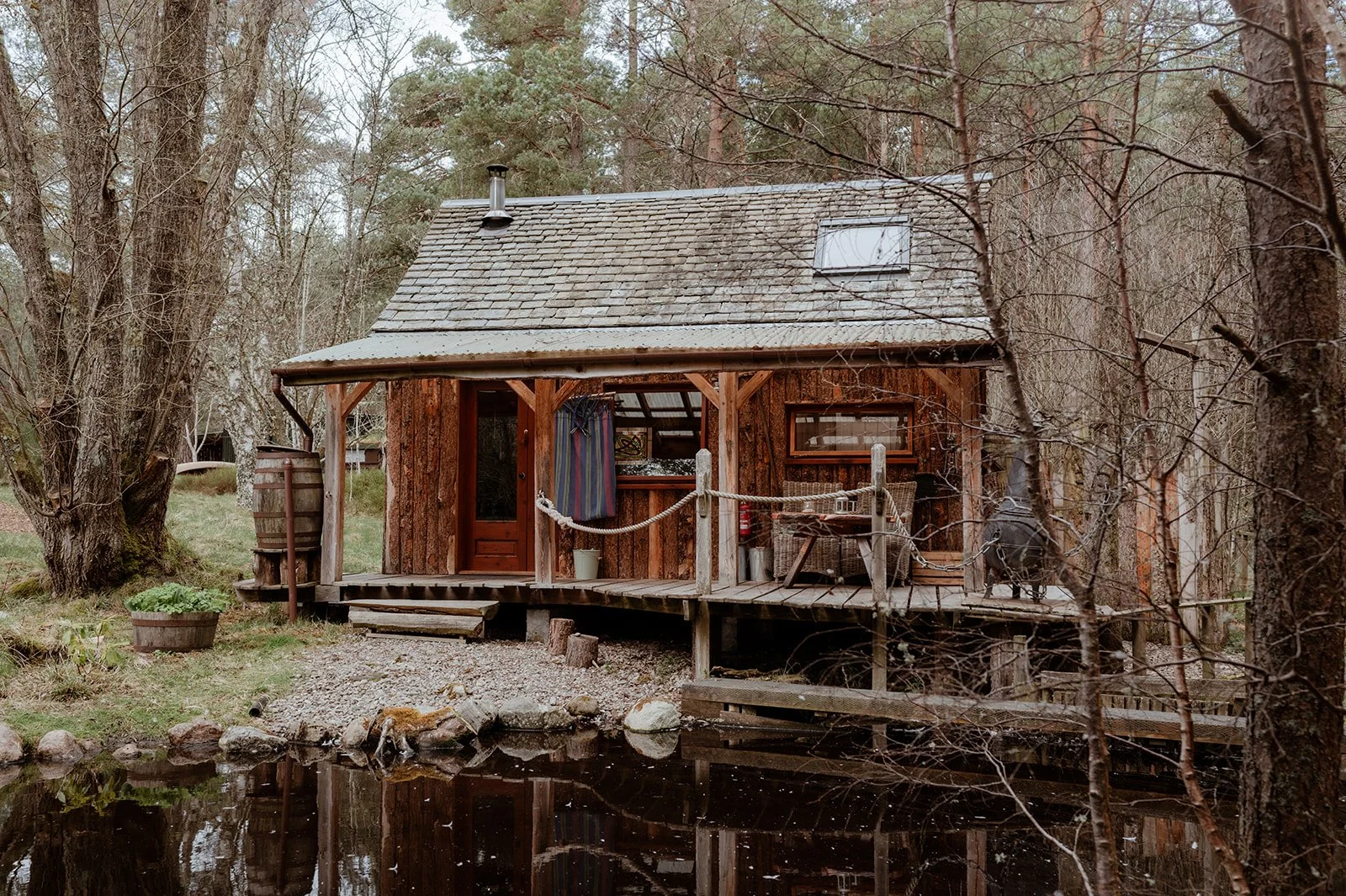 A rustic wooden cabin on stilts surrounded by trees, next to a pond with a rocky edge.