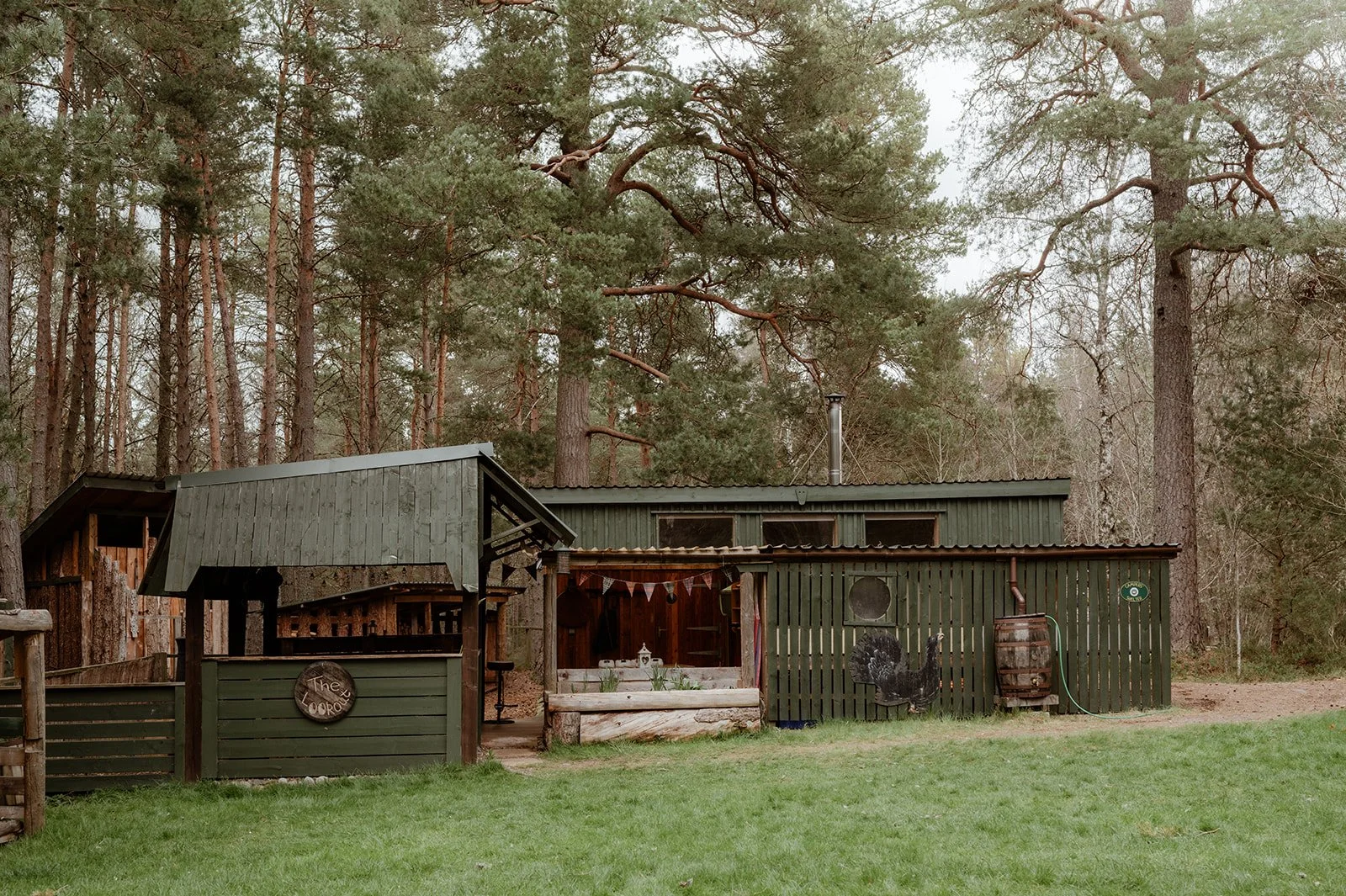 An off-grid bunkhouse in the cairngorms national park. The structure is green and surrounded by pine forest with a grass field in the foreground.