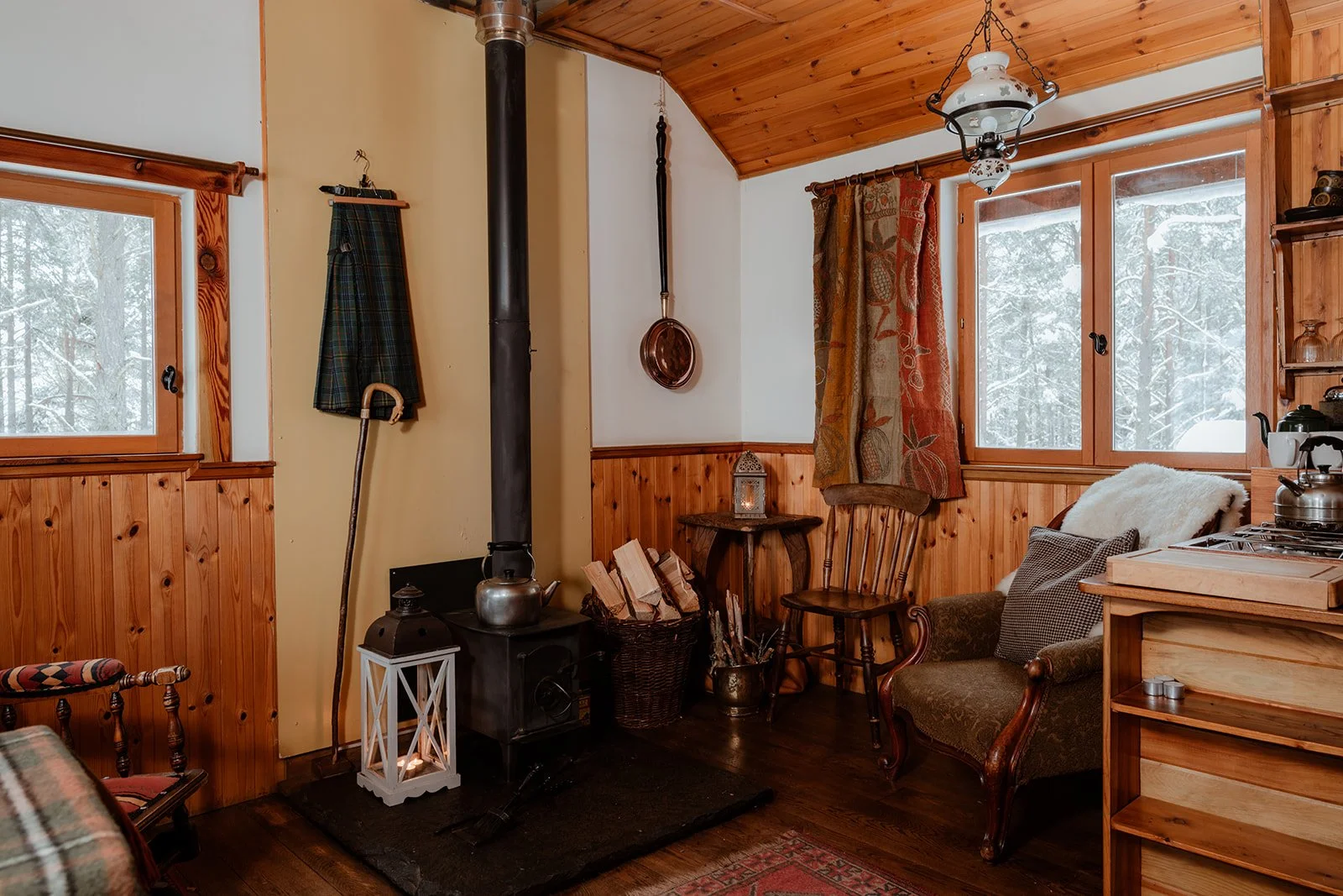 Cozy cabin interior with wood-paneled walls, a wood stove, and furniture including chairs, a small table, and a vintage armchair, with windows showing snow outside.