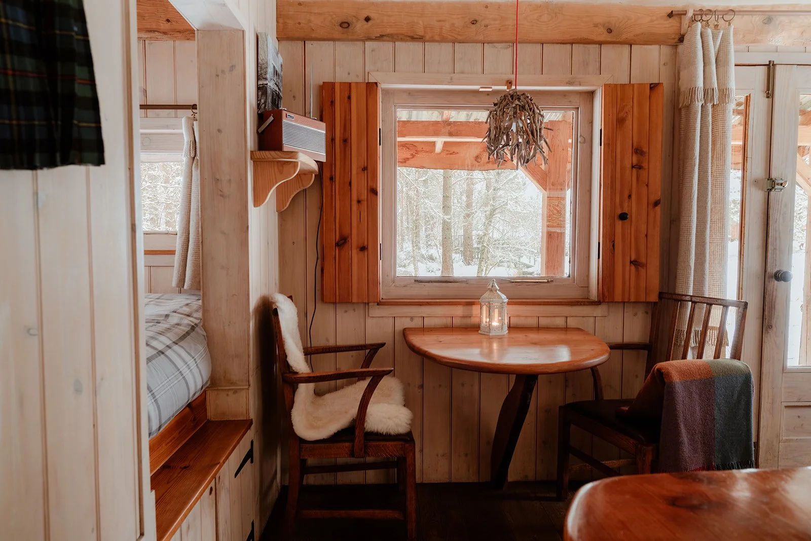 Cozy wooden cabin interior with a round dining table, two chairs, window view of snowy trees, and a built-in bed partially visible on the left.