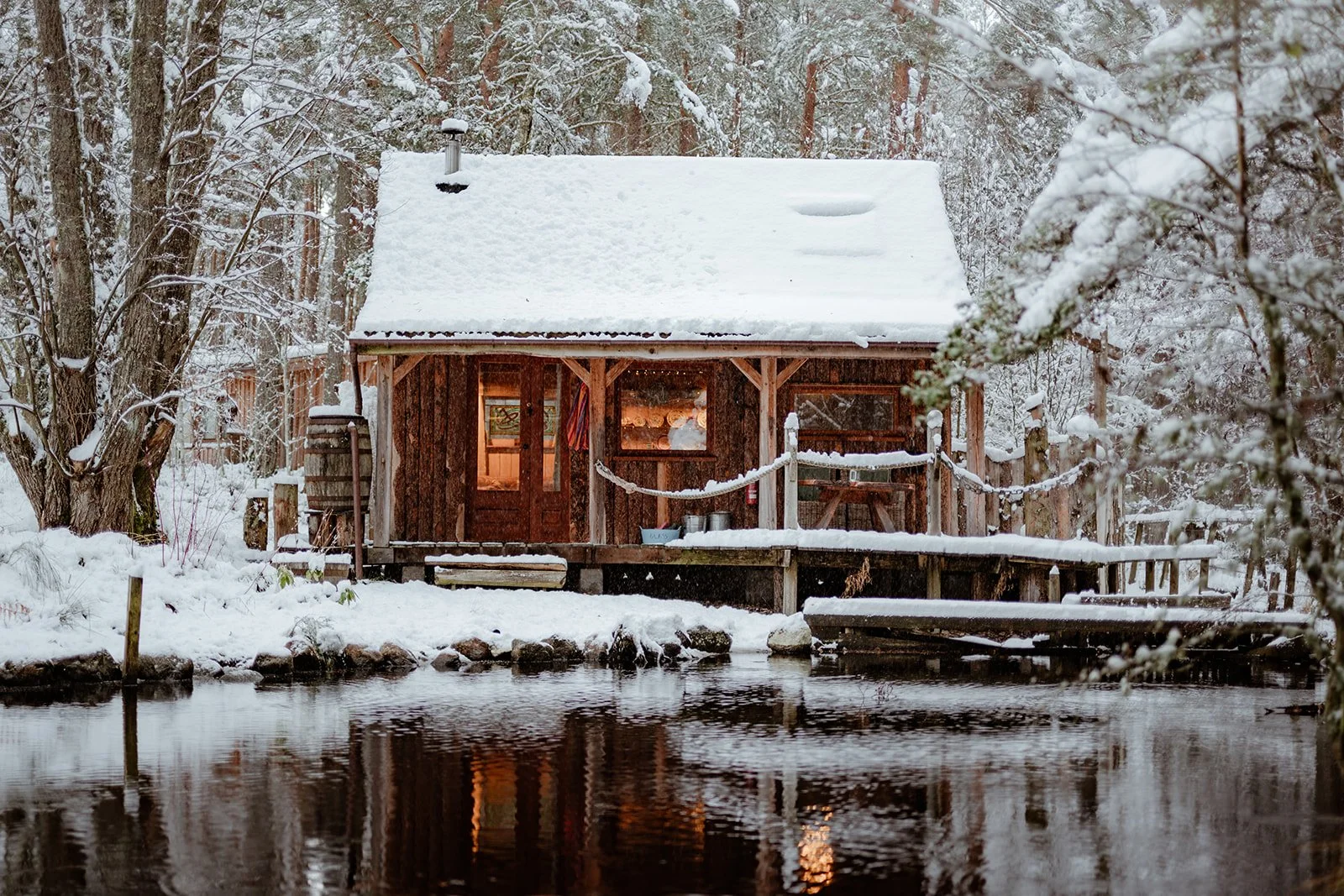 A snowy cabin by a river with a snow-covered roof, trees around, and warm lights inside.