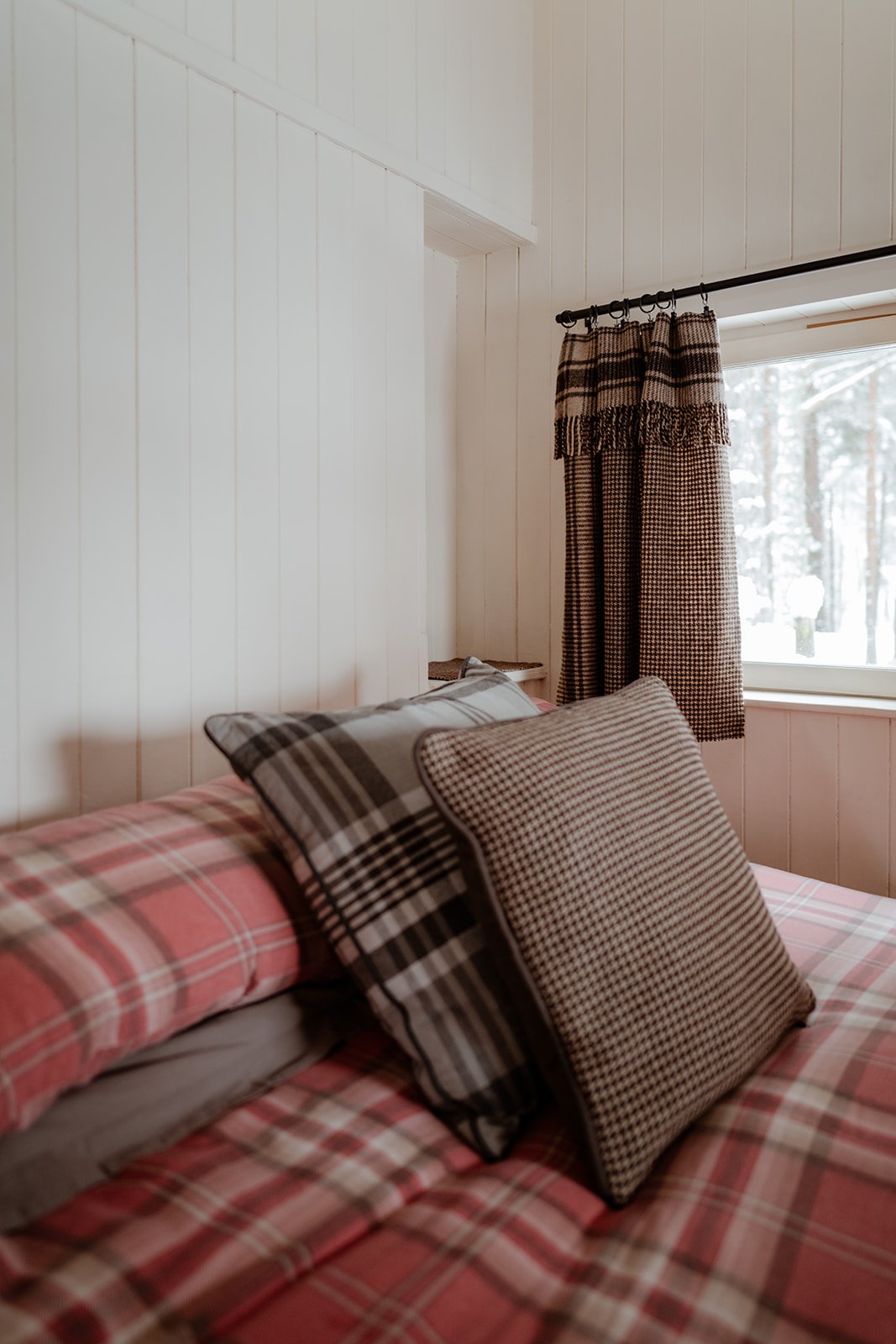 A cozy bedroom corner with a bed covered in red plaid bedding, topped with two pillows featuring checkered patterns. A window with a view of a snowy outdoor scene has a dark curtain with a houndstooth pattern.