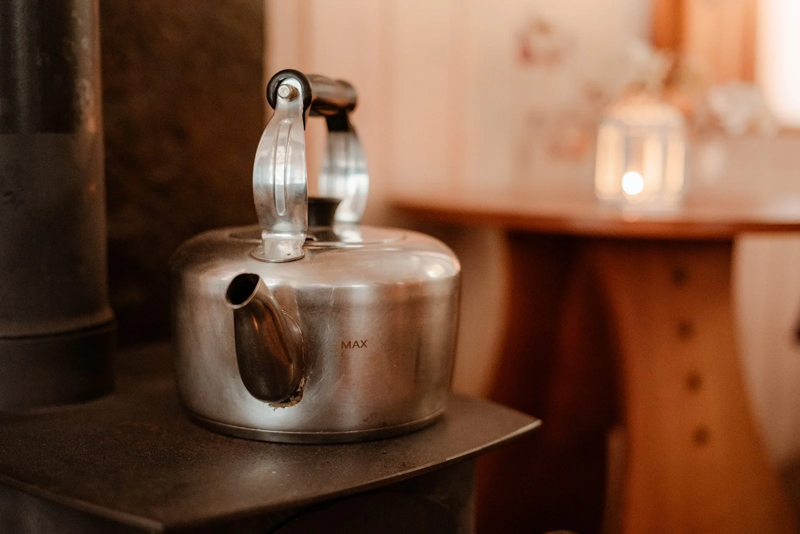 A stainless steel kettle on a stove with a wooden table and a blurred background in a cozy kitchen or dining area.