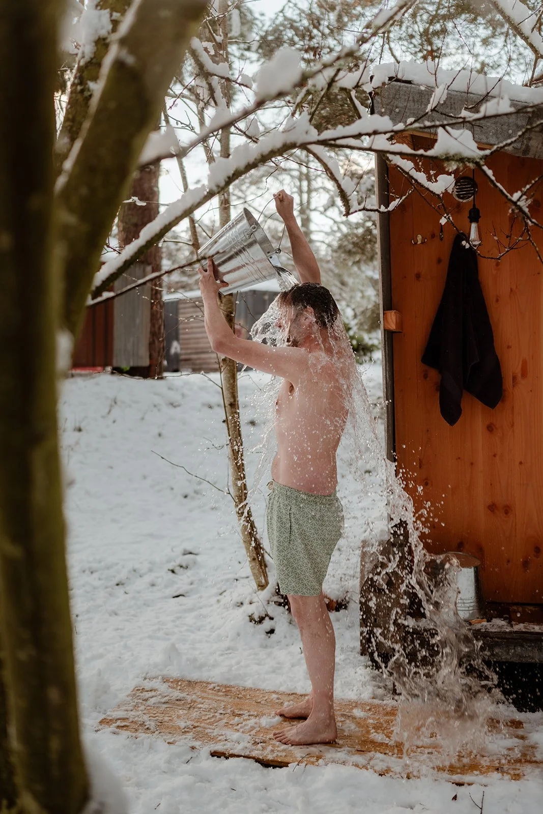 A young man in shorts pouring water over his head outside on a snowy day, standing on a wooden platform near a small wooden shed with snow-covered trees around.