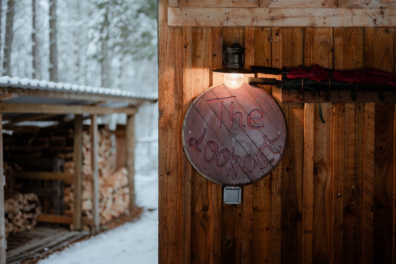 A wooden sign with the words 'The Outpost' written on it, mounted on a wooden wall near an outdoor woodpile with snow in the background.