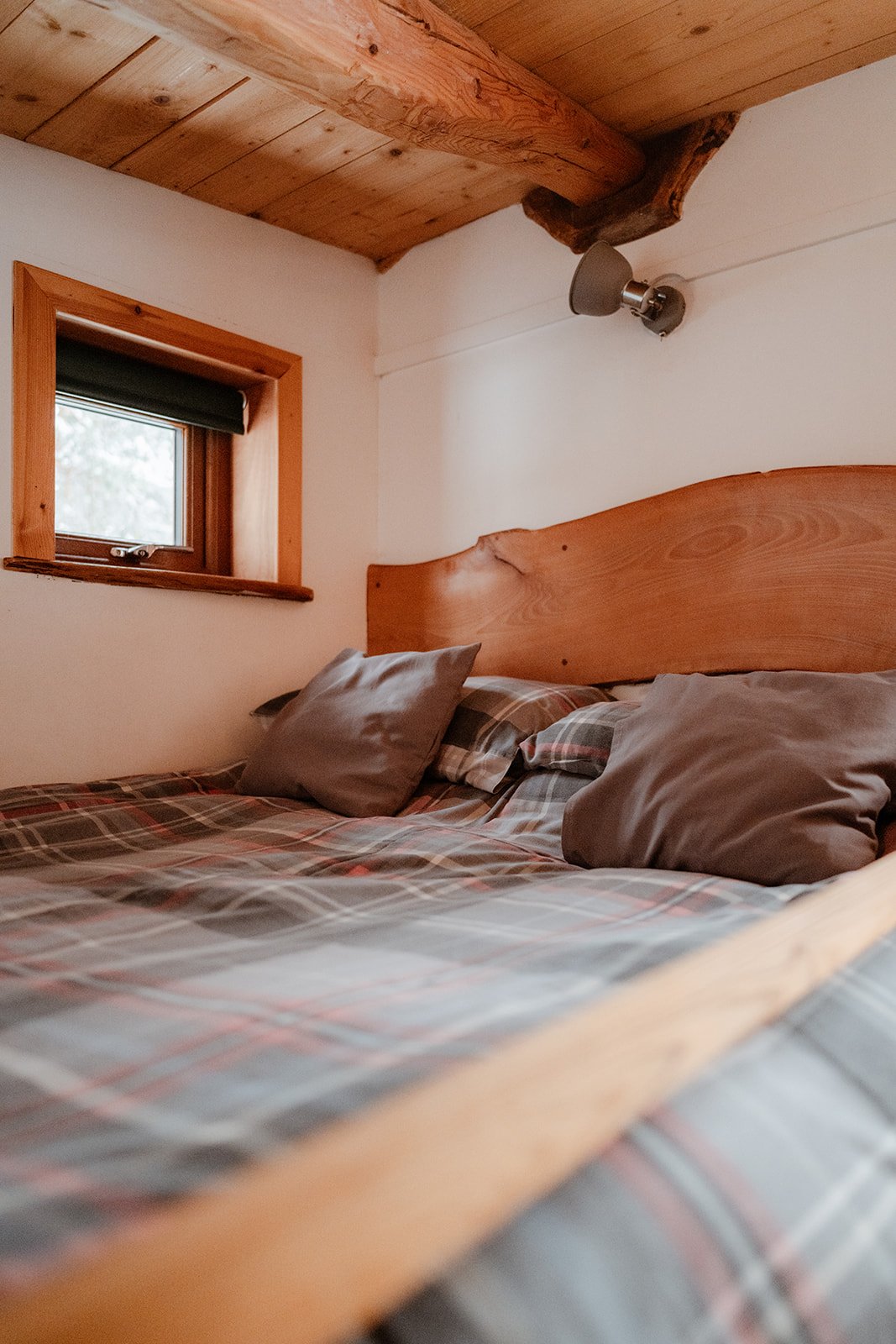 A cozy bedroom with a wooden bed frame, plaid bedding, and pillows. There is a small window with a black shade and a wall-mounted reading lamp, all with wooden accents and a wooden ceiling.