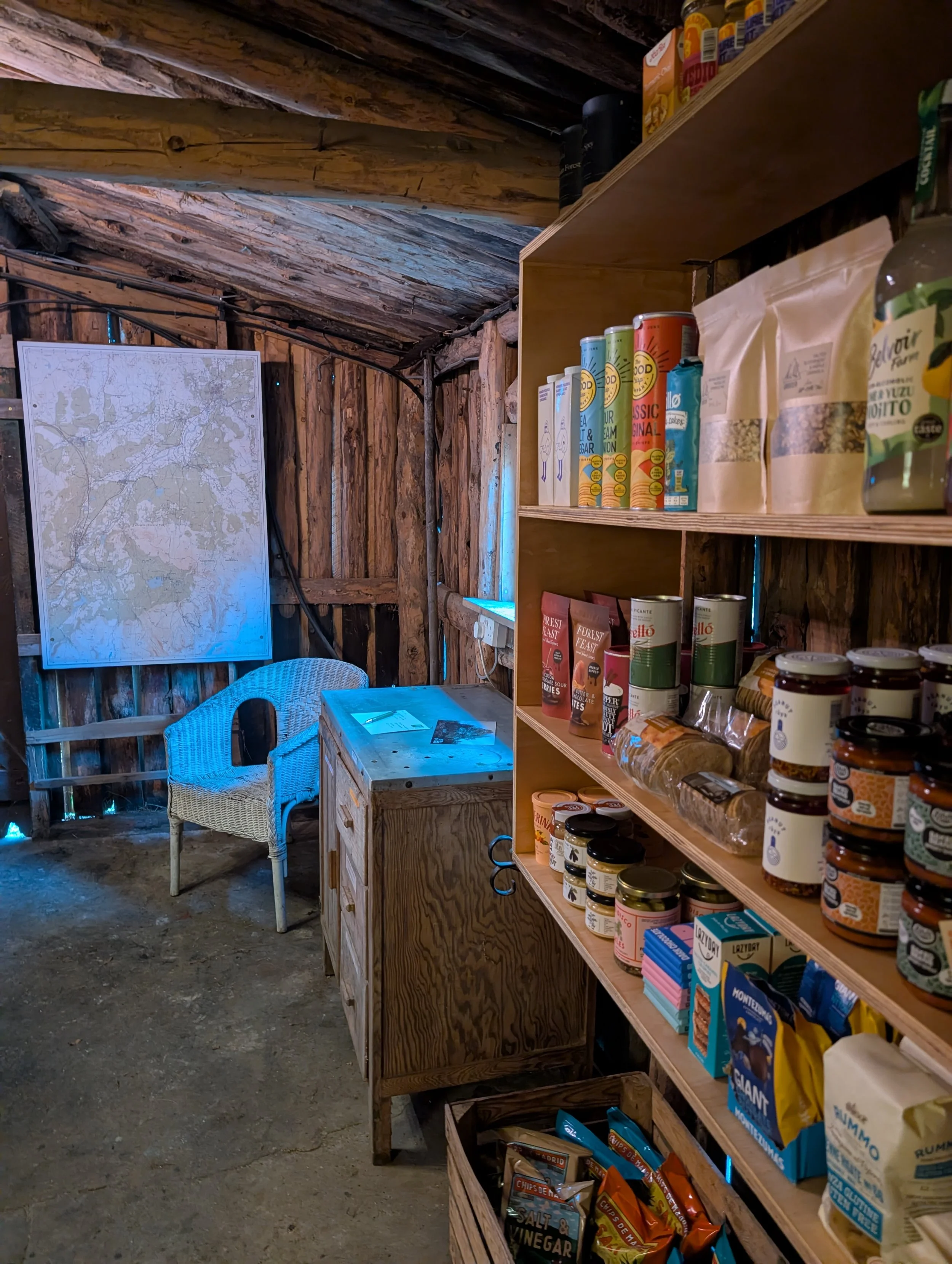 A cozy rustic pantry with wooden walls and ceiling, featuring a wooden shelf stocked with snacks, canned goods, and beverages, a small wooden cabinet, a wicker chair, and a large map on the wall.