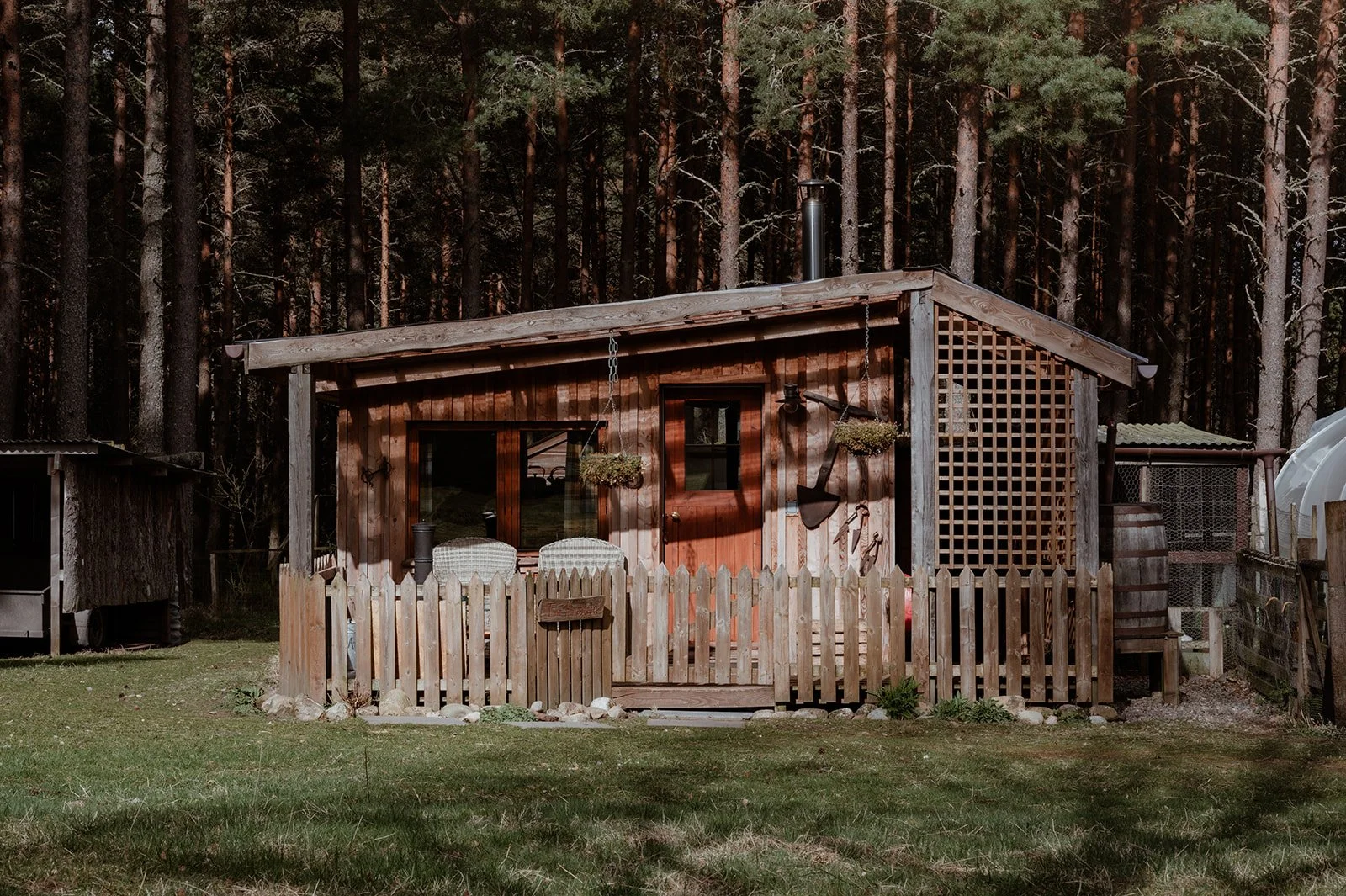 Small wooden cabin with a fenced front yard, surrounded by tall trees in a forest setting.