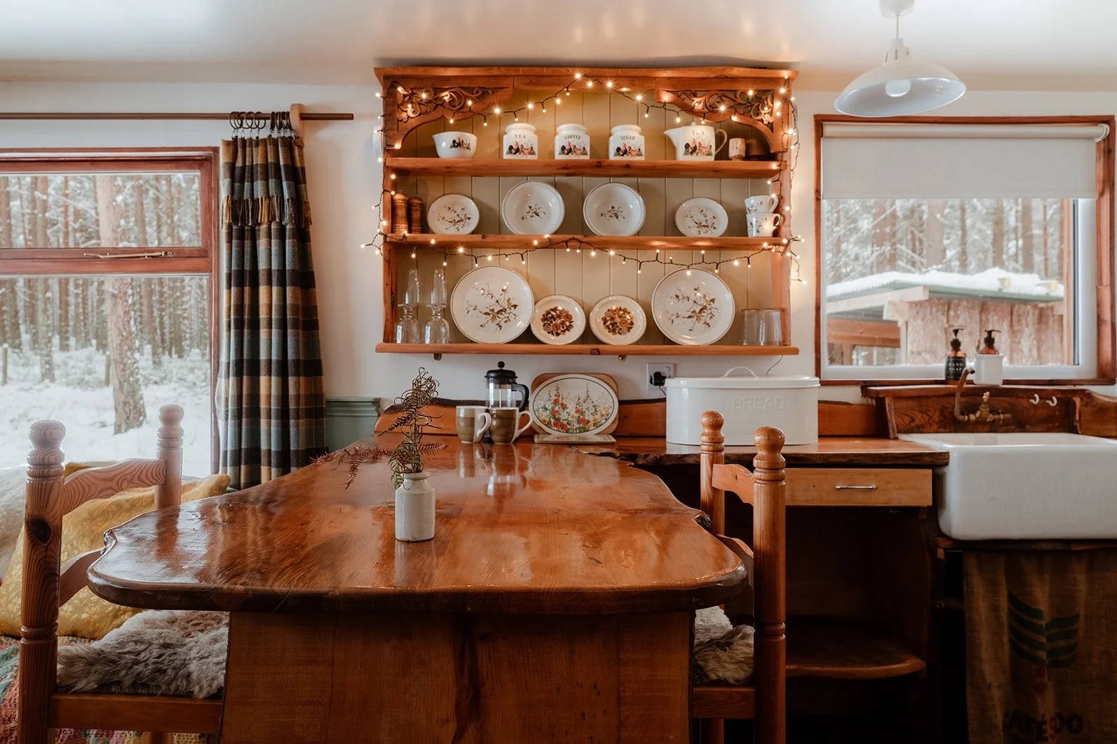 Cozy kitchen with wooden dining table and open shelving displaying vintage dishes, set against windows showing a snowy outdoor landscape.