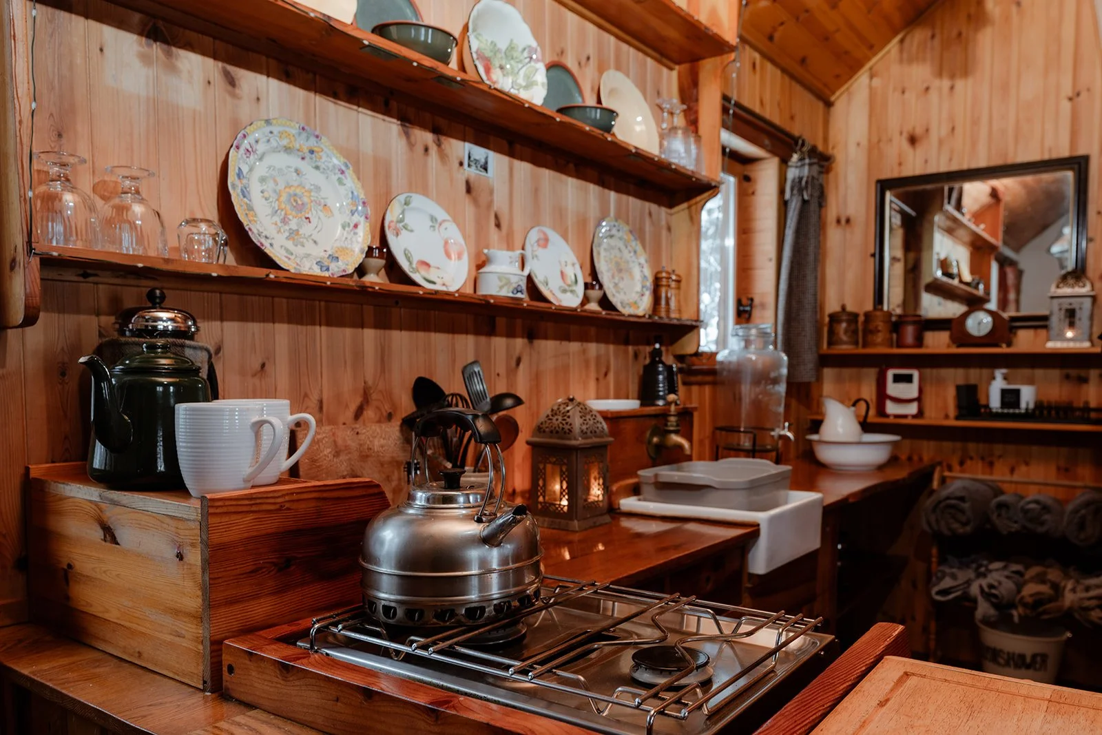 A cozy kitchen with wooden walls and shelves displaying decorative plates, glassware, and kitchen utensils. A kettle and mugs sit on a stove, with a small lantern and window in the background.