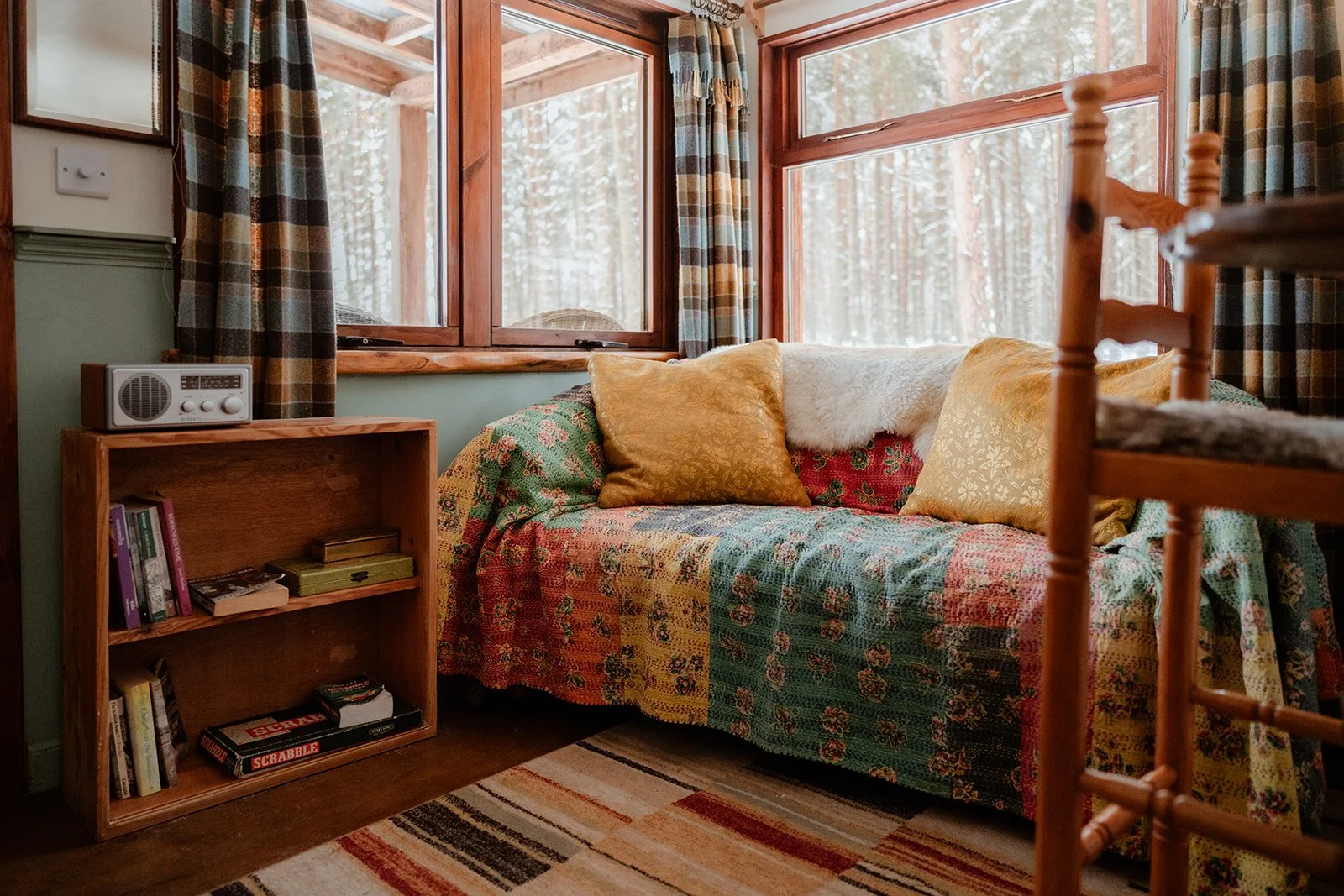 Cozy sitting area in a cabin with a wooden framed sofa covered by a colorful quilt, decorated with yellow and white pillows, beside a small bookshelf with books, under a window showing a snowy forest outside, with plaid curtains and a small radio on 