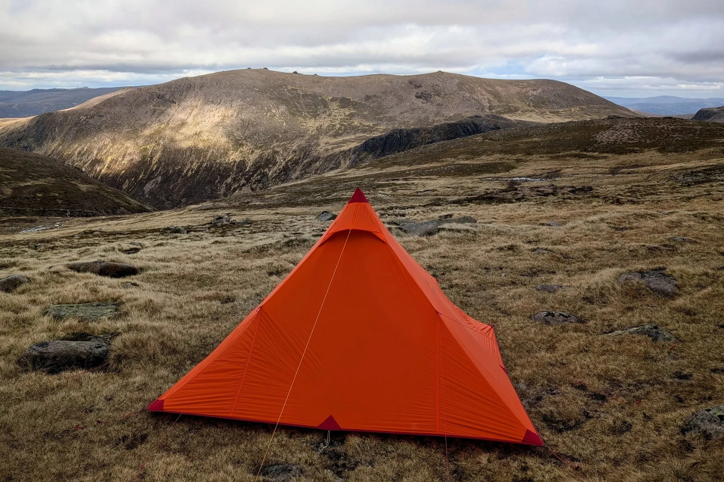 An orange tent wildcamping in the mountains with big hills in the background. anda cloudy sky in the cairngorms national park