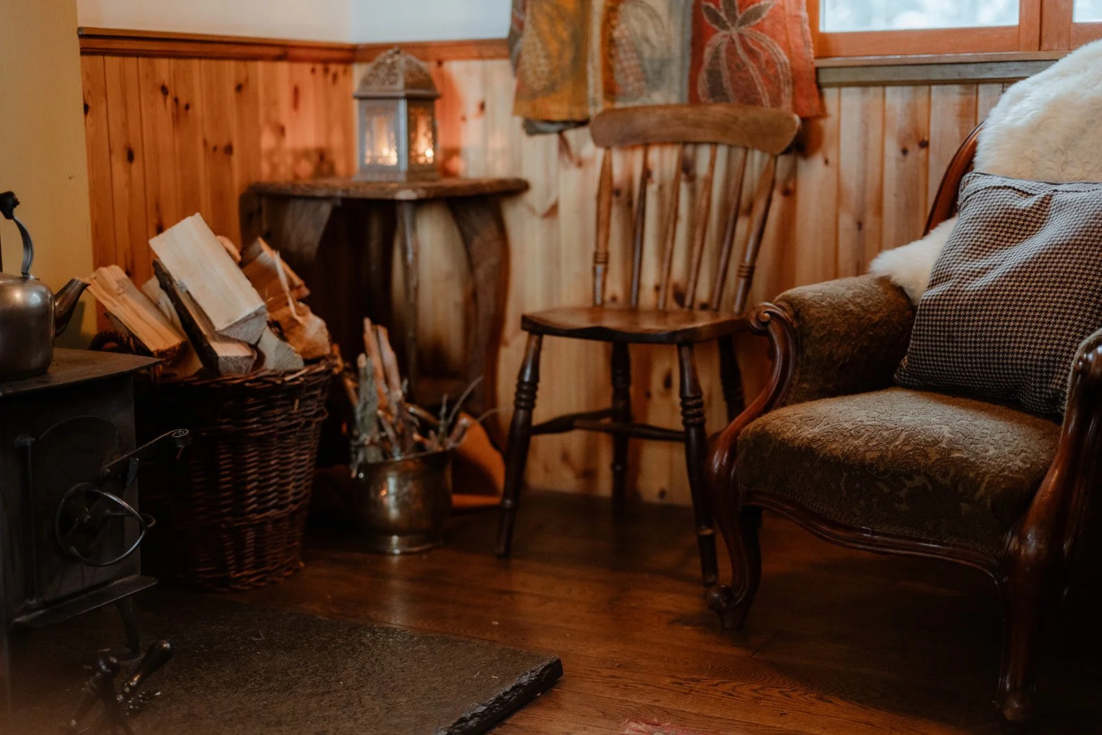 Cozy cabin interior with wood-paneled walls, a vintage armchair with cushions, a wooden chair, a basket of firewood, and a lantern on a small table.