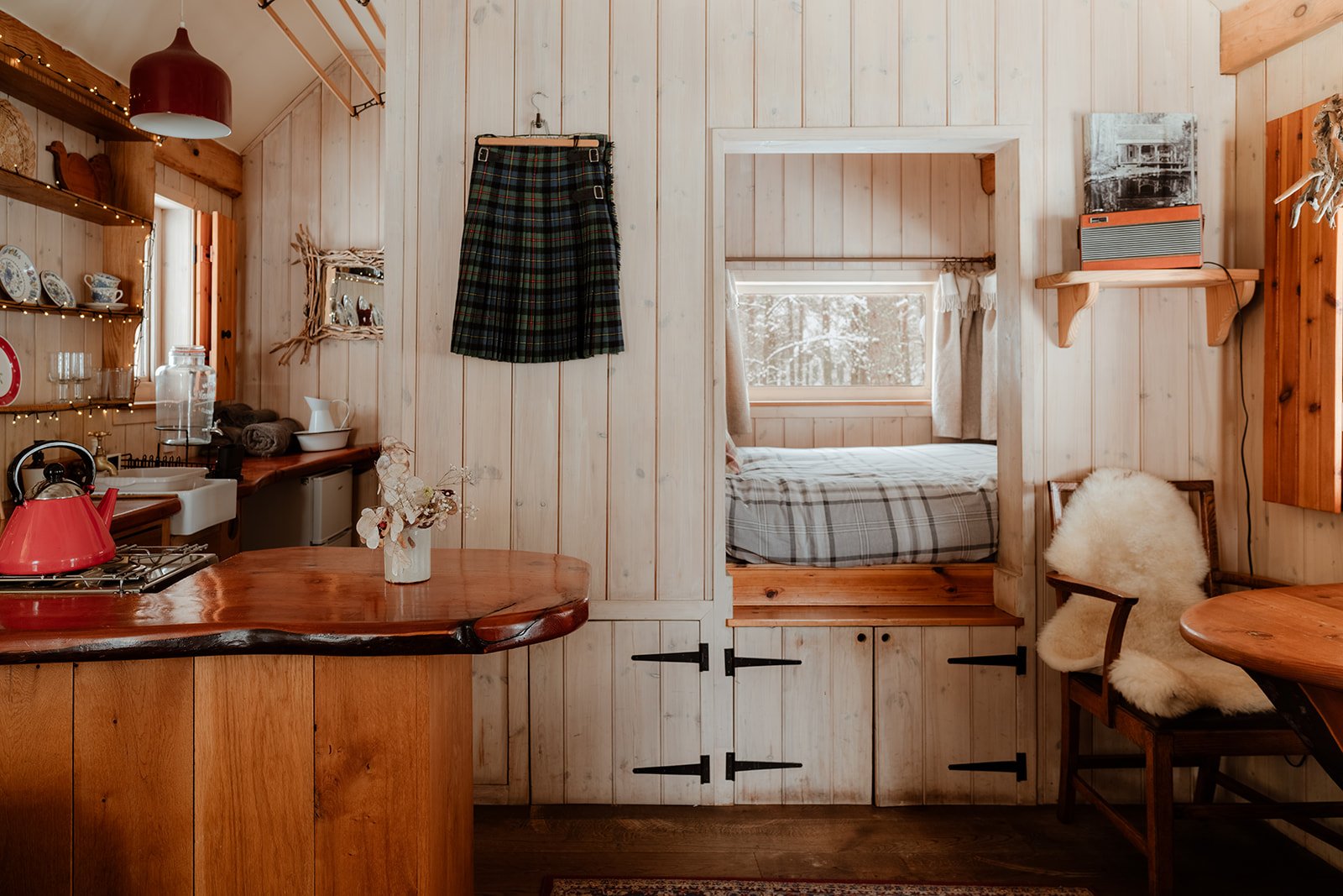Wood-paneled interior of a cozy room with a small kitchen area, a plaid skirt hanging on the wall, a doorway leading to a bedroom with a window, a sheepskin chair, and shelves with decorations.