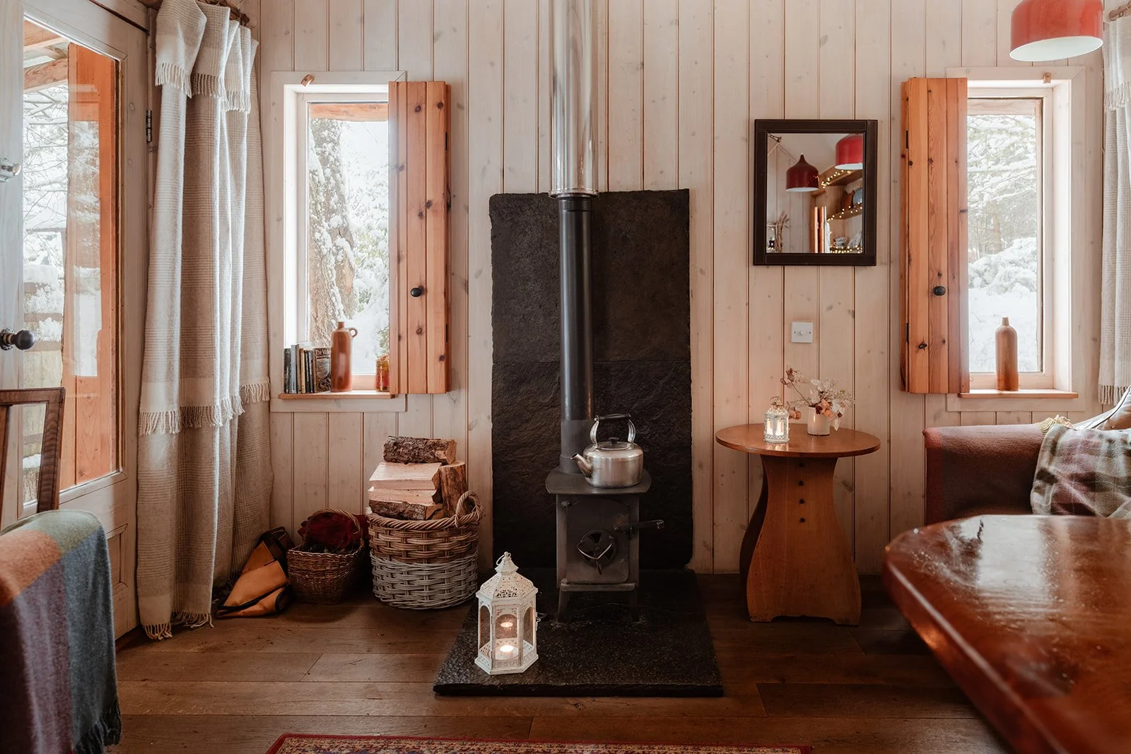 A cozy living room with light wood-paneled walls, two windows with wooden shutters and vases, a wood stove with a kettle, a small table with flowers, and a lantern on the floor.