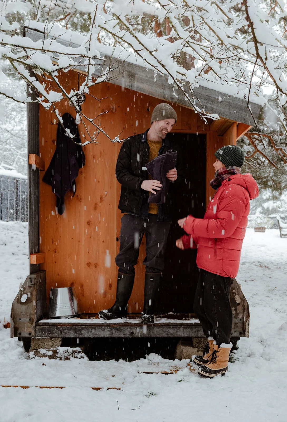 A man and a woman stand on a small wooden lookout in a snowy outdoor setting, smiling and talking. The man is holding a jacket, and both are dressed warmly with hats and winter gear.