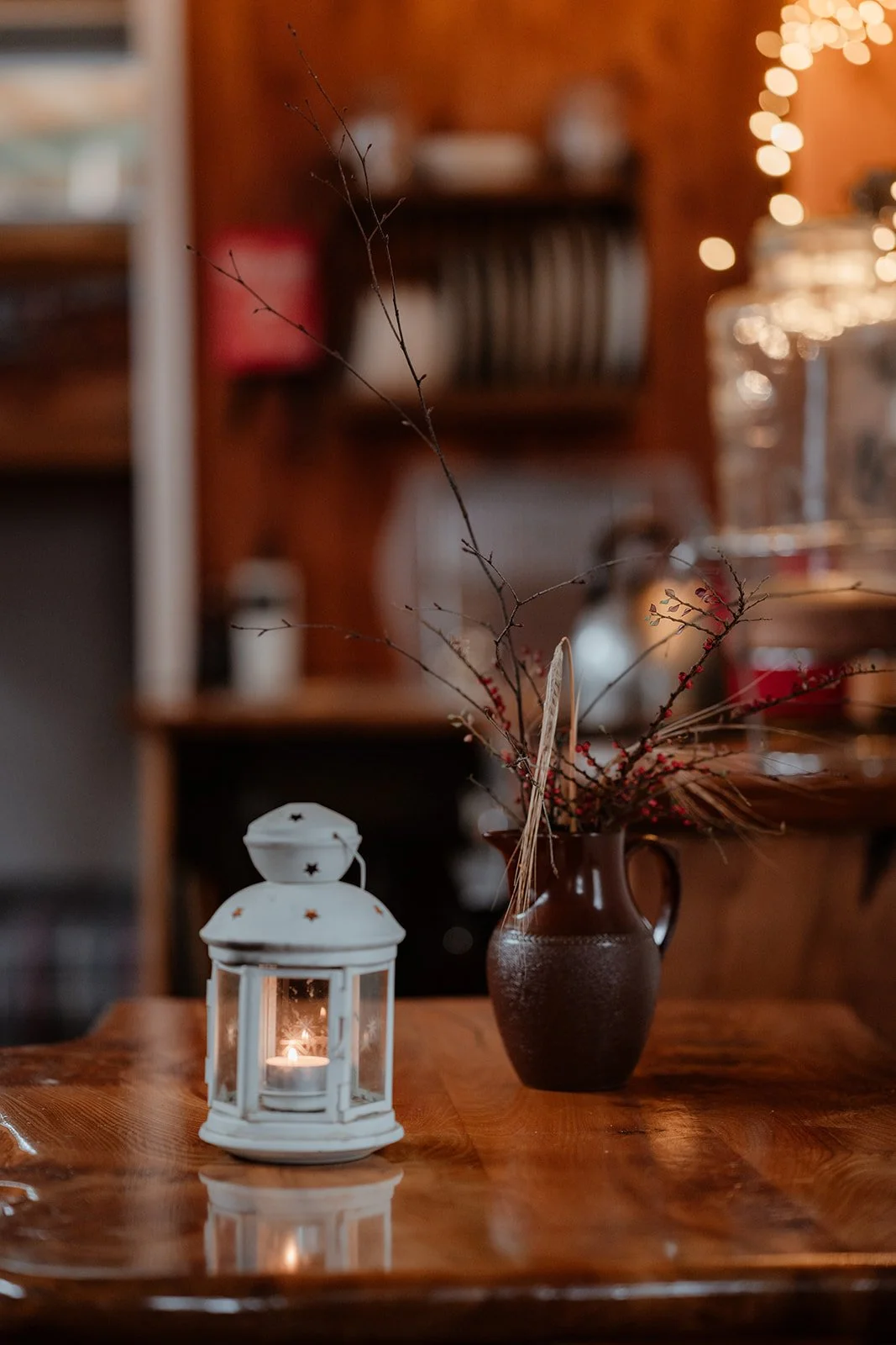 A lit white lantern and a brown vase with twigs and red berries on a wooden table in a cozy, warmly lit room.