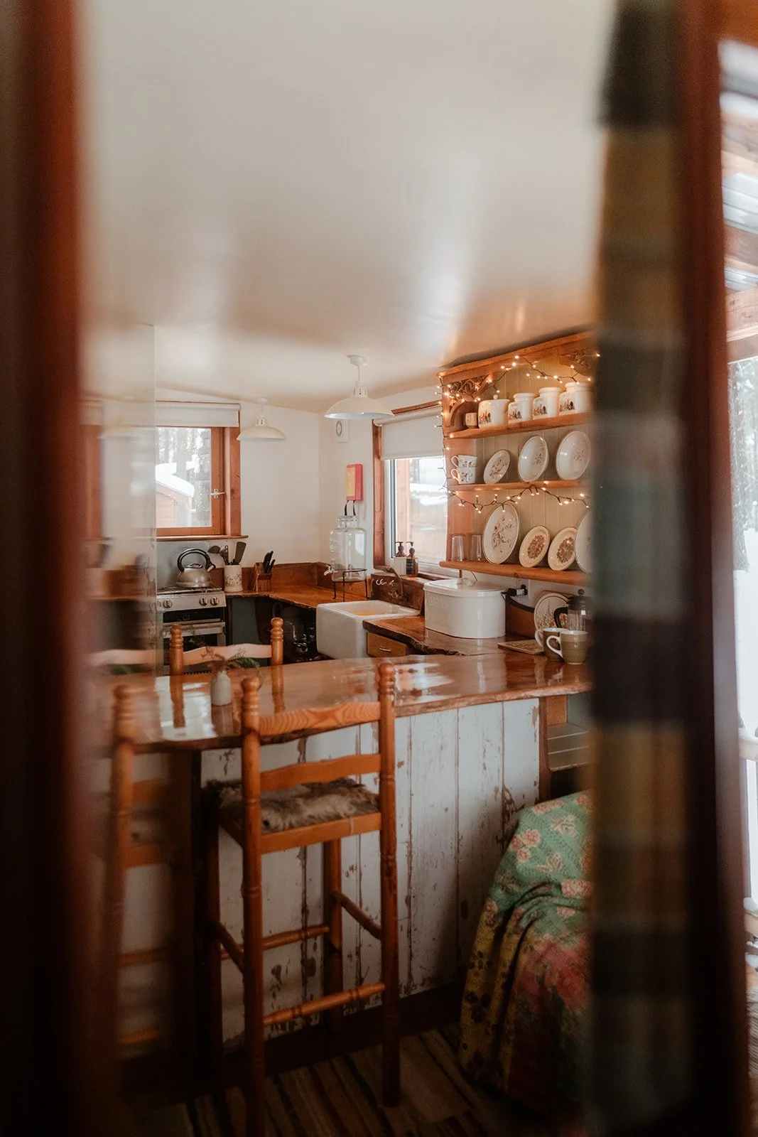 A cozy kitchen viewed through a doorway with wooden framing, featuring wooden chairs, a wood counter, white wall cabinets, and open shelving displaying plates, mugs, and decorative items, with string lights adding warm ambiance.