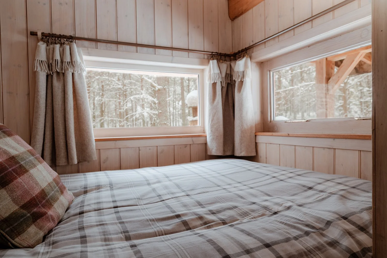 Cozy bedroom with wooden paneled walls, a large bed with plaid bedding and a plaid pillow, and two windows showing a snowy forest outside.