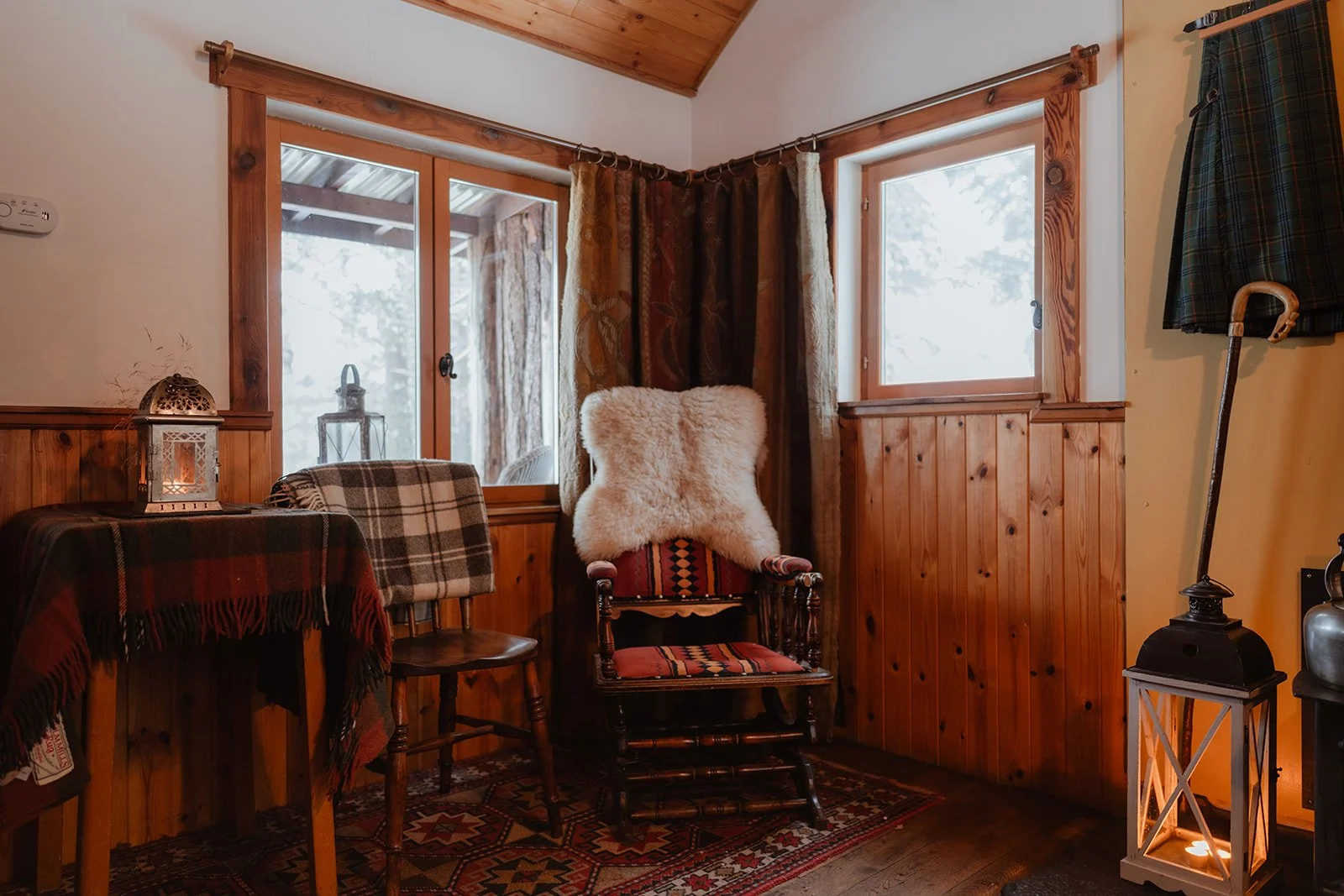 Cozy corner of rustic cabin with wood-paneled walls, frosted windows, a rocking chair with a fluffy throw pillow, a small table with a lantern, and a plaid shawl draped over a chair.