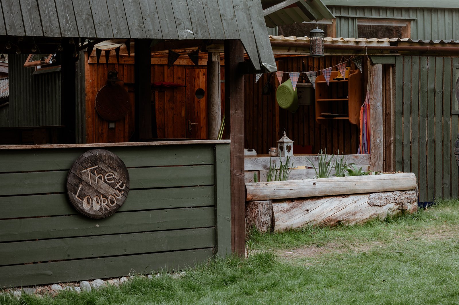 Wooden outdoor structure with a sign reading "The Lodge" on a green fence, a raised garden bed with green plants, and decorative items like lanterns and bunting.