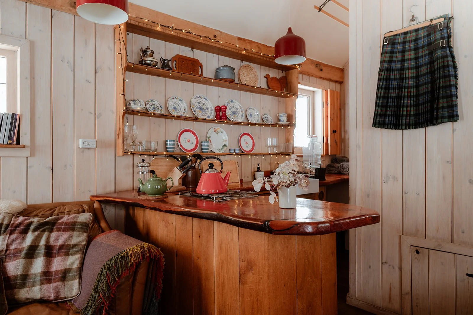 A cozy kitchen with wooden walls and shelves displaying decorative plates, teapots, and jars, decorated with string lights. There is a wooden counter with a green teapot, red kettle, and a vase with artificial flowers. A tartan skirt hangs on the wal