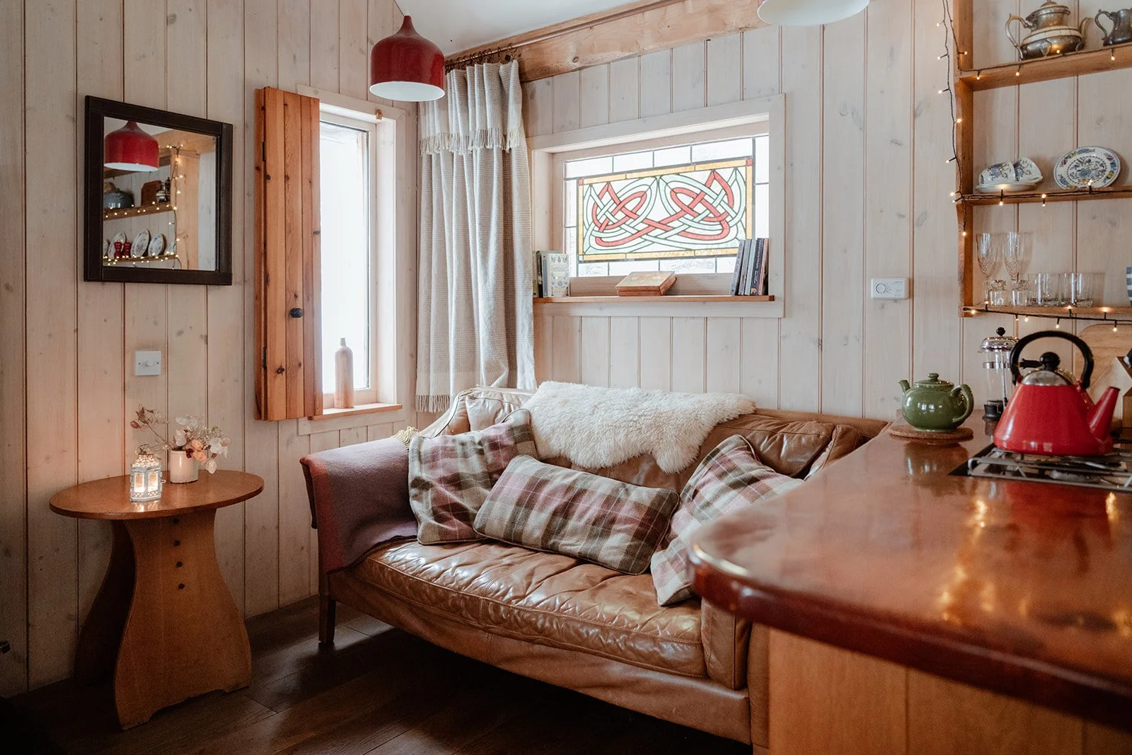 Cozy living room with wooden paneled walls, a leather couch with tartan pillows and a throw blanket, a small round wooden side table with candles and flowers, windows with curtains and a stained glass window, and open shelves with dishes and glasswar