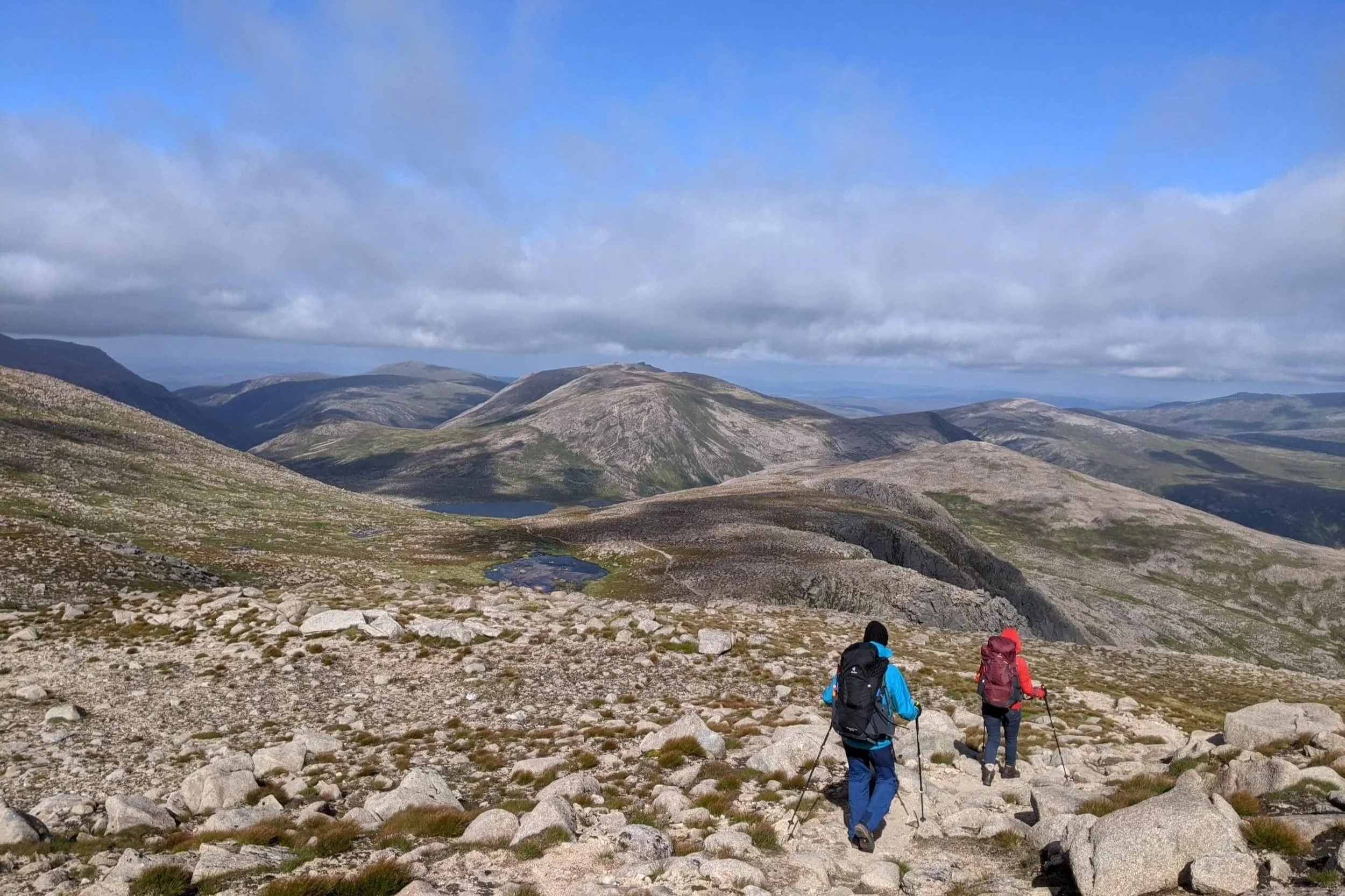 two hikers walking down a mountain path towards a small lake. You can see mountains in the distance with clouds low and a blue skie above.