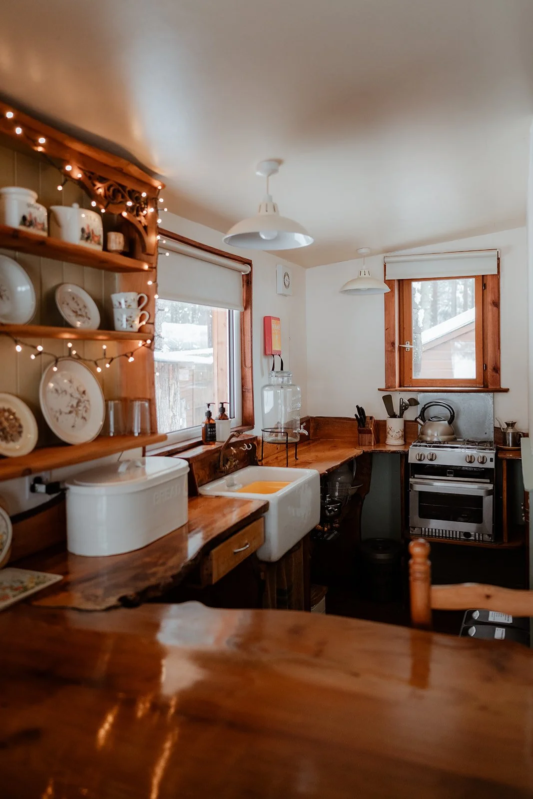 A cozy kitchen with wooden countertops, a white farmhouse sink, a window with a blind, a stove with a kettle, shelves with dishes, and warm lighting.