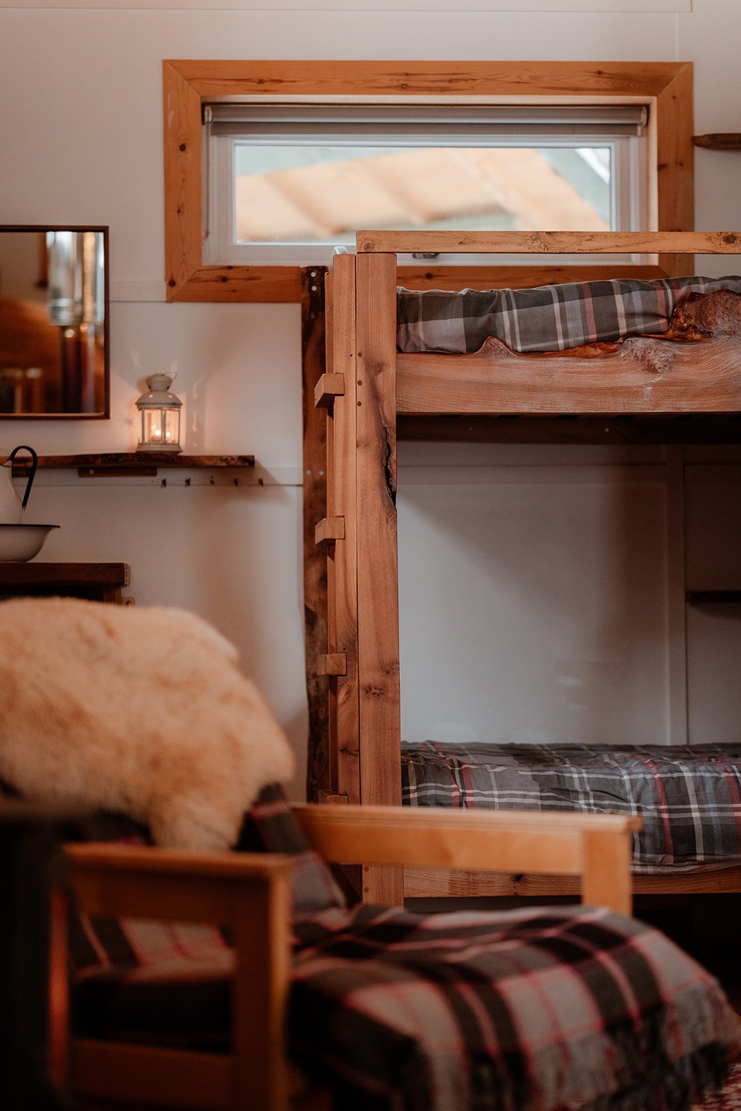 A cozy rustic bedroom with a wooden bunk bed, plaid bedding, a window with wooden trim, and a lantern on a wall shelf.