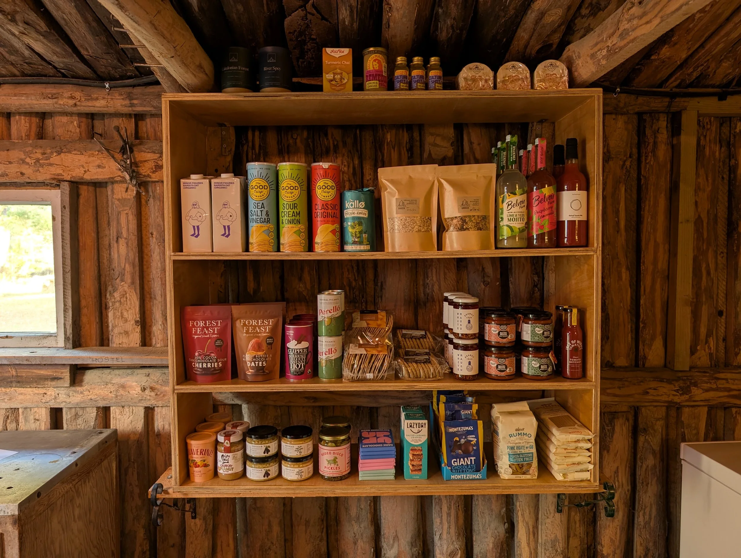 Wooden shelf stocked with various packaged food items including snacks, sauces, and canned goods inside a rustic wooden room.