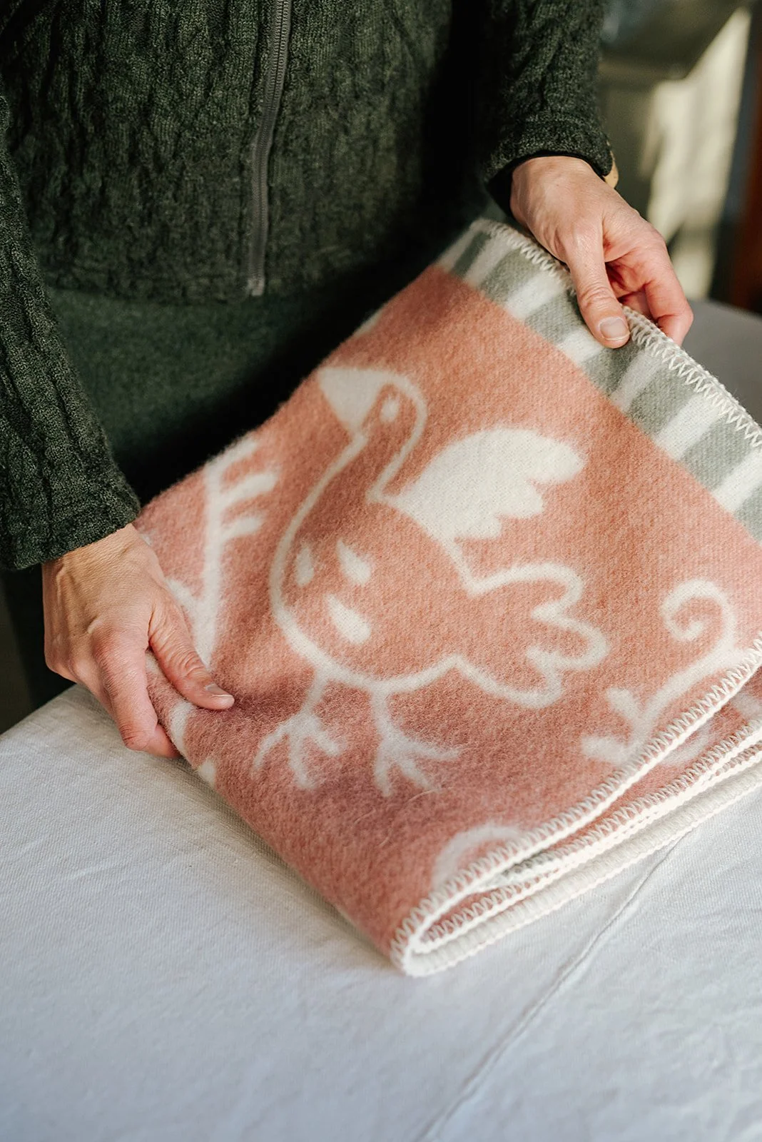 Person folding a pink and white patterned wool blanket with a swan design at Lilla Ullboden i Kumla.