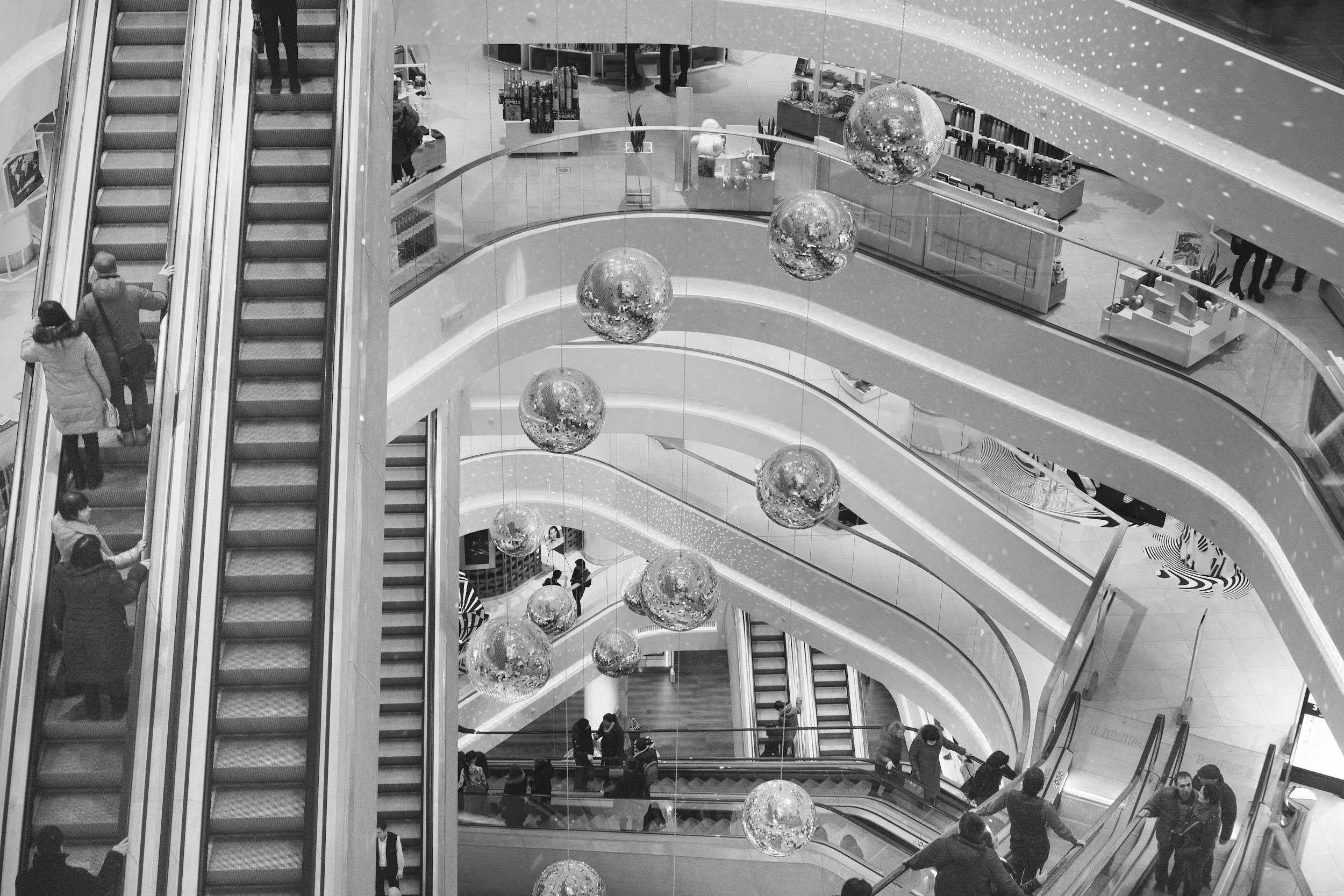 Multiple levels of a shopping mall with stores, escalators, and shoppers walking around, including a Mango clothing store and other retail shops.
