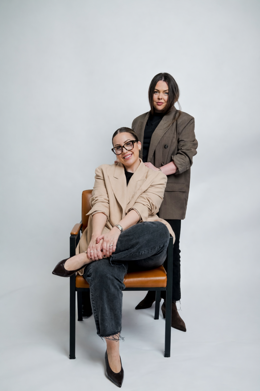 Two women in professional attire pose together against a plain white background, with one seated in a chair and the other standing behind her.