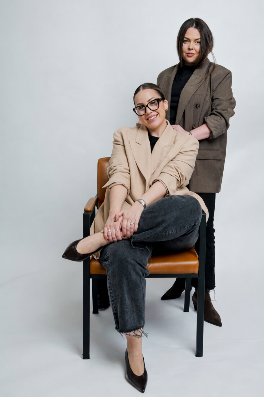 Two women posing together in a studio with a plain background. One is sitting in a chair with legs crossed, wearing glasses, a beige blazer, and black jeans. The other is standing behind her, resting her hand on the seated woman's shoulder, wearing a brown blazer and black pants.