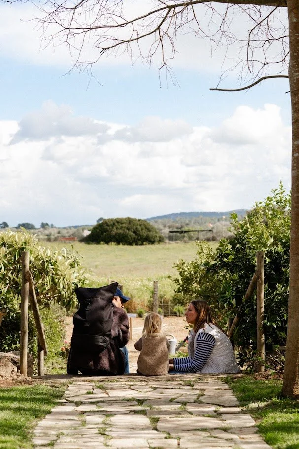 Grupo de cuatro personas, incluyendo dos niños, sentados en un camino de piedra en un entorno rural con árboles y campo abierto. Discutiendo o conversando.